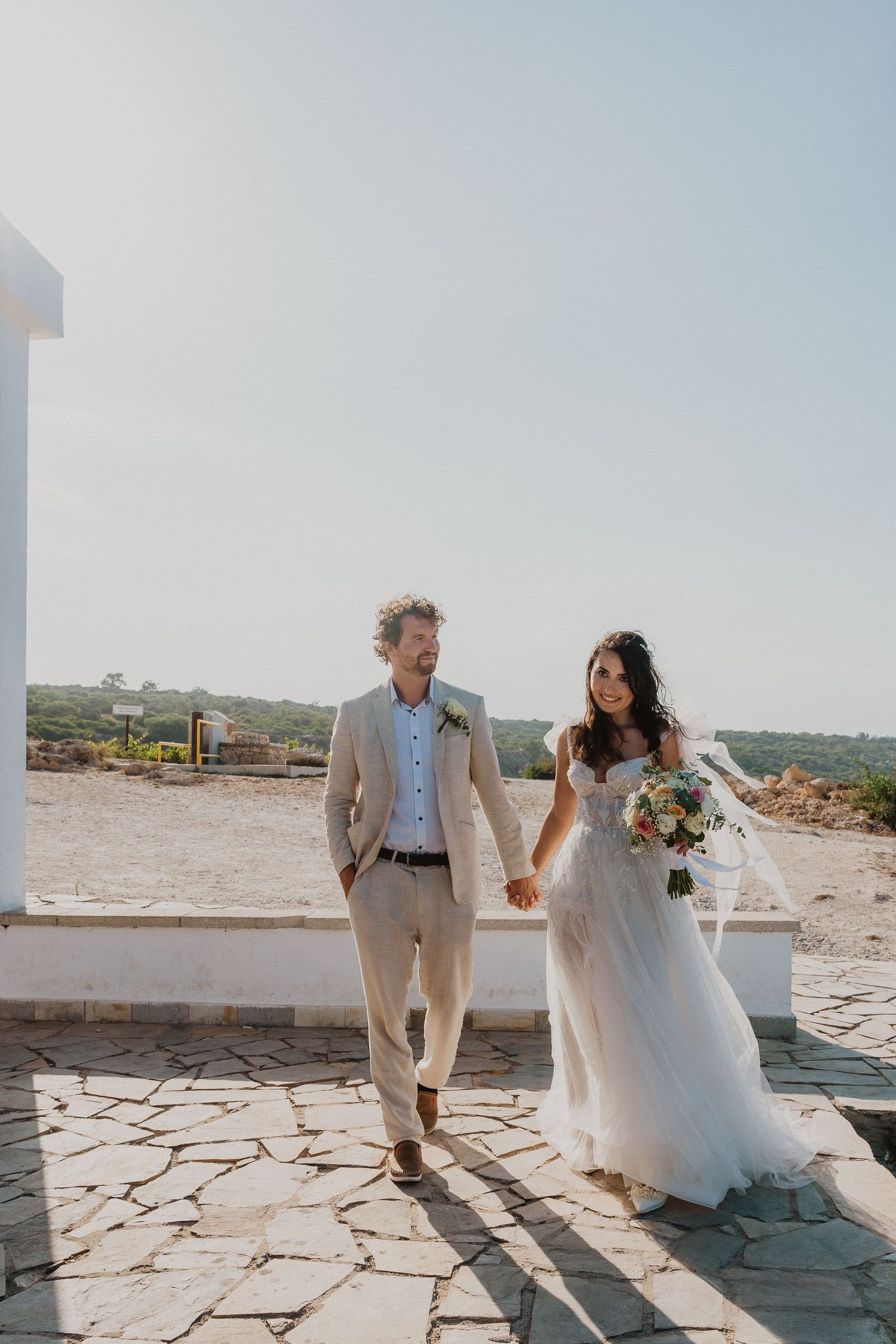 Newlyweds stroll together on a Mediterranean terrace with scenic views in the background.