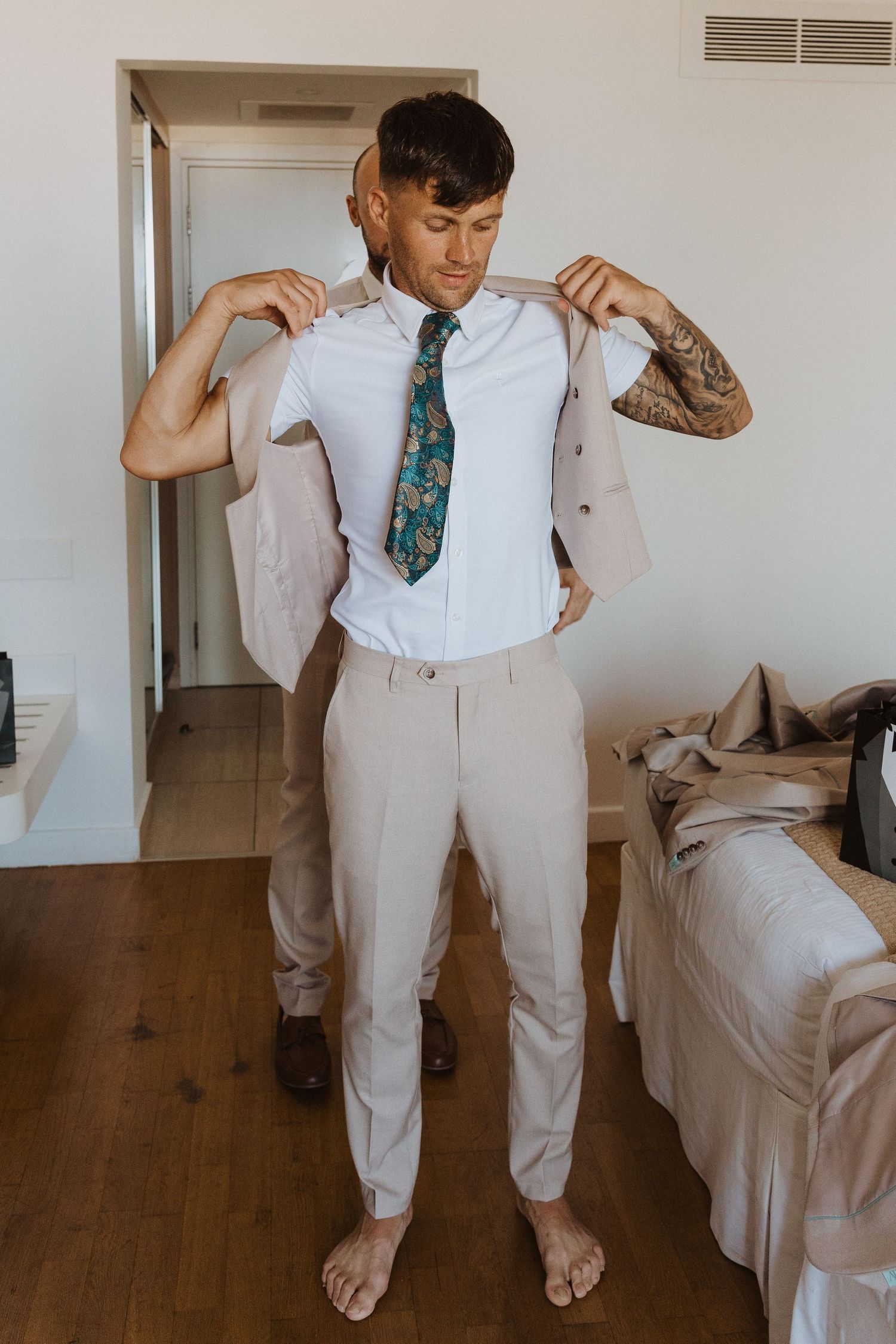 A person getting dressed in a light beige suit and patterned tie stands barefoot on hardwood floors in a bedroom.