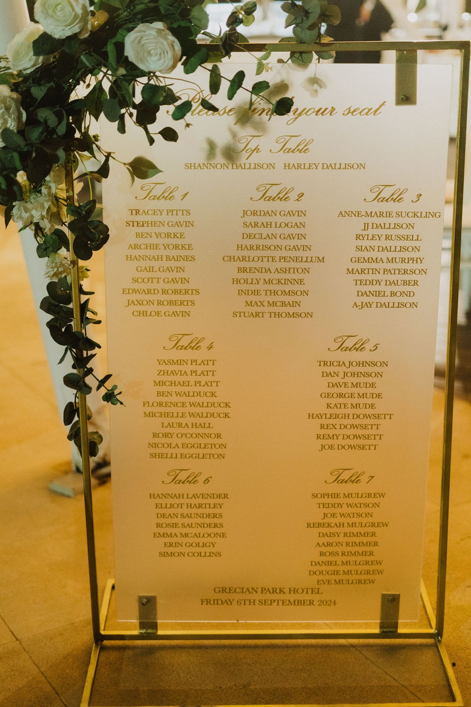Elegant wedding seating chart display with floral greenery decoration against golden background.