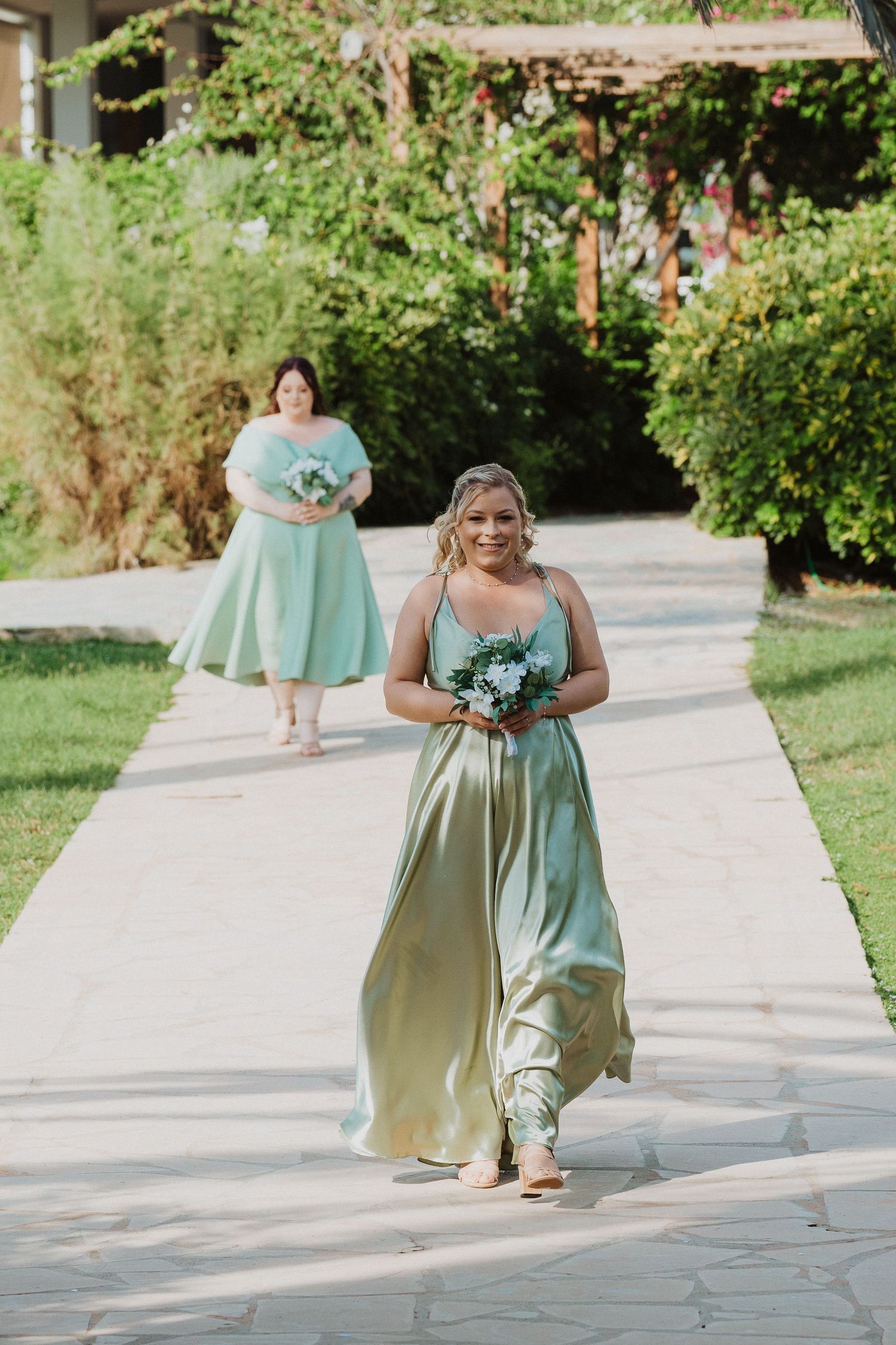 Bridesmaid in mint green flowing dress walks down outdoor garden path holding blue bouquet.