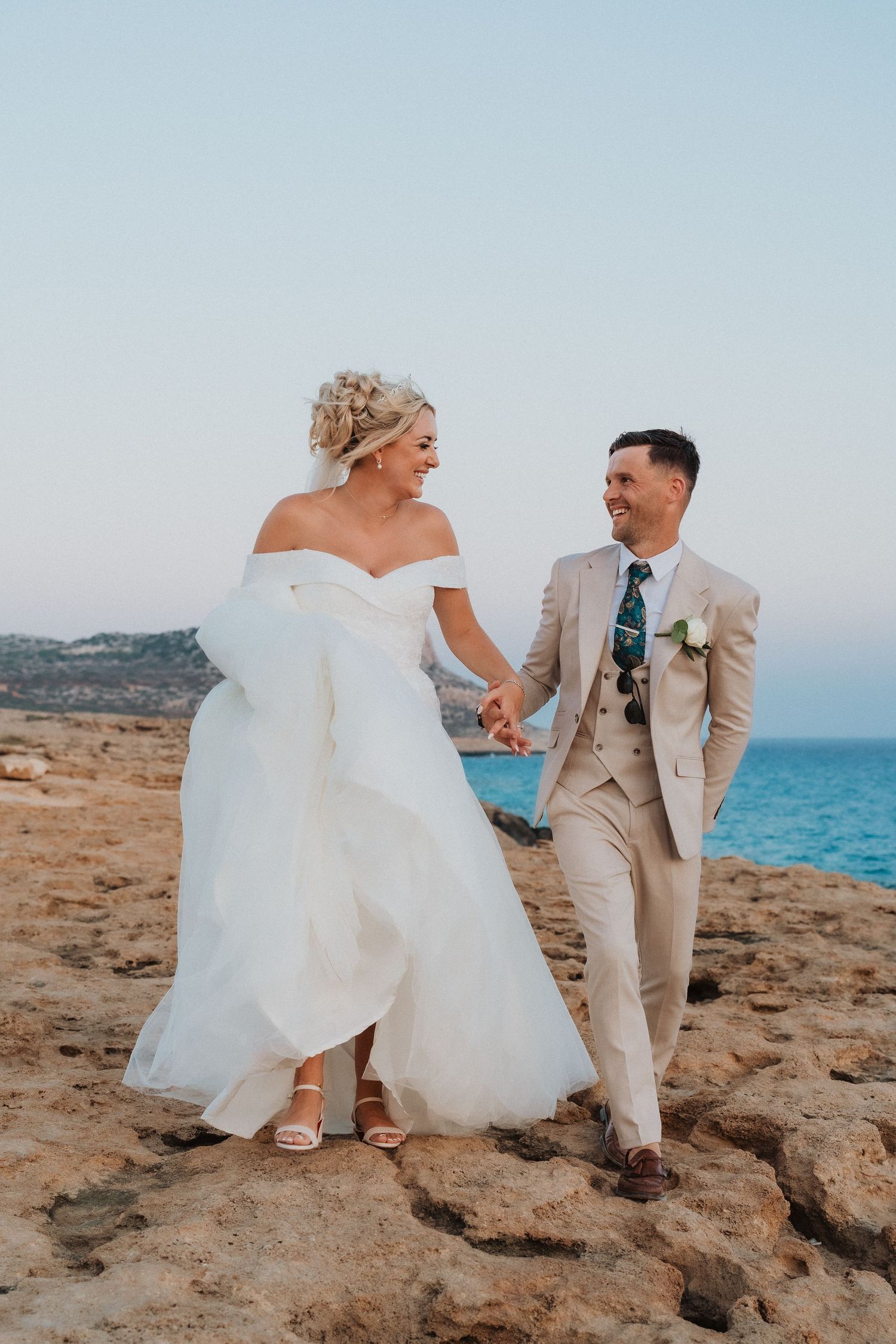 A couple shares a laugh while walking along scenic coastal cliffs in wedding attire.