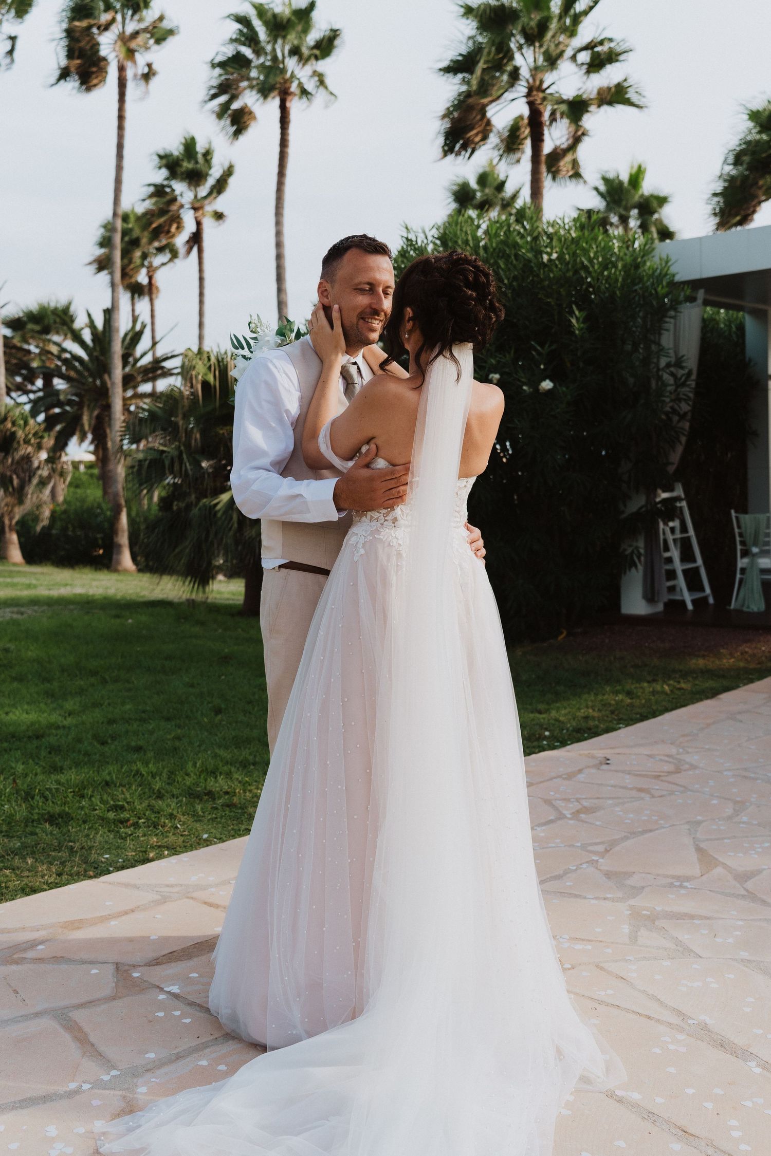 A couple shares an intimate moment on their wedding day at an outdoor venue with tall palm trees in the background.