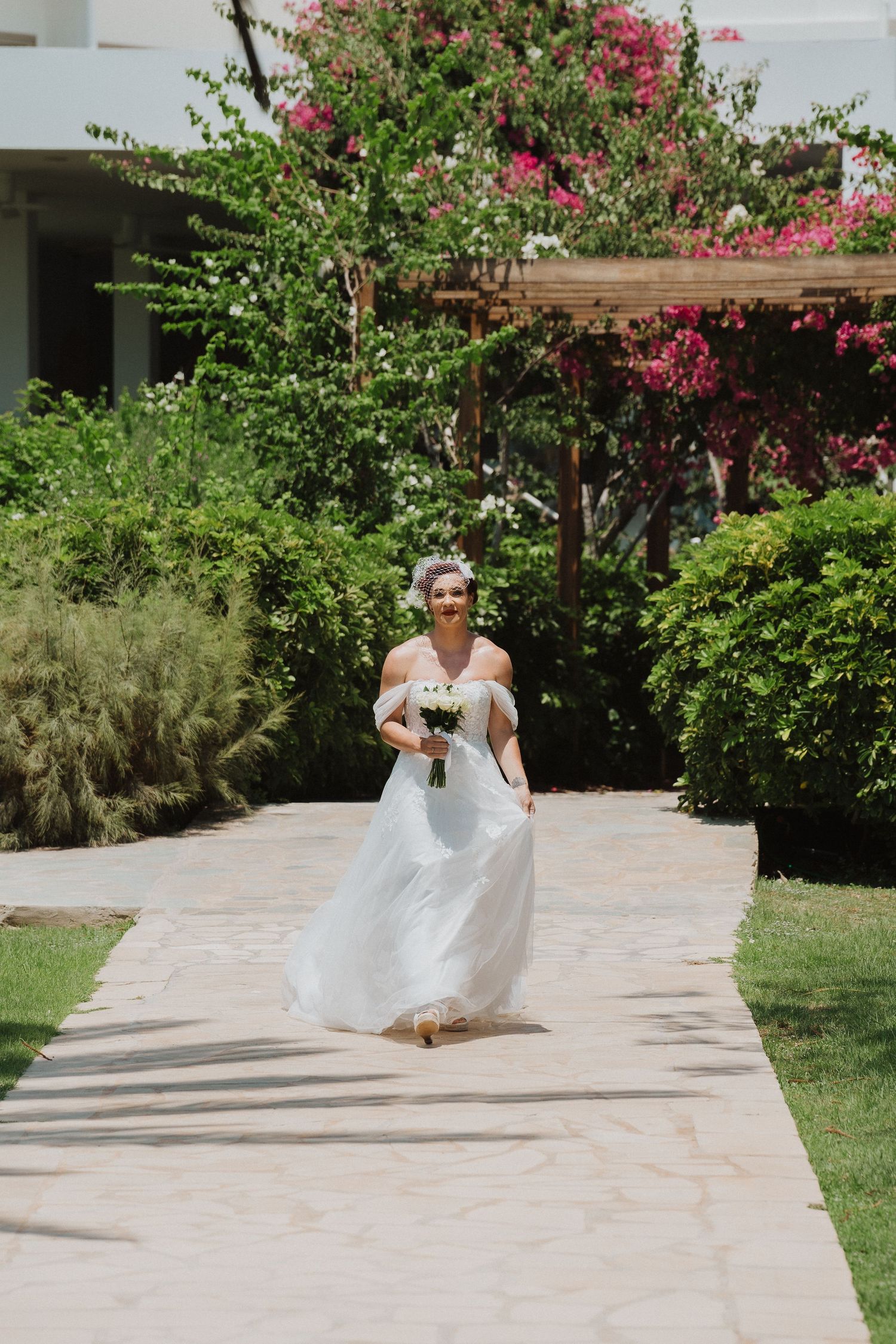 A bride in an off-shoulder white gown walks along a garden path with pink bougainvillea flowers.