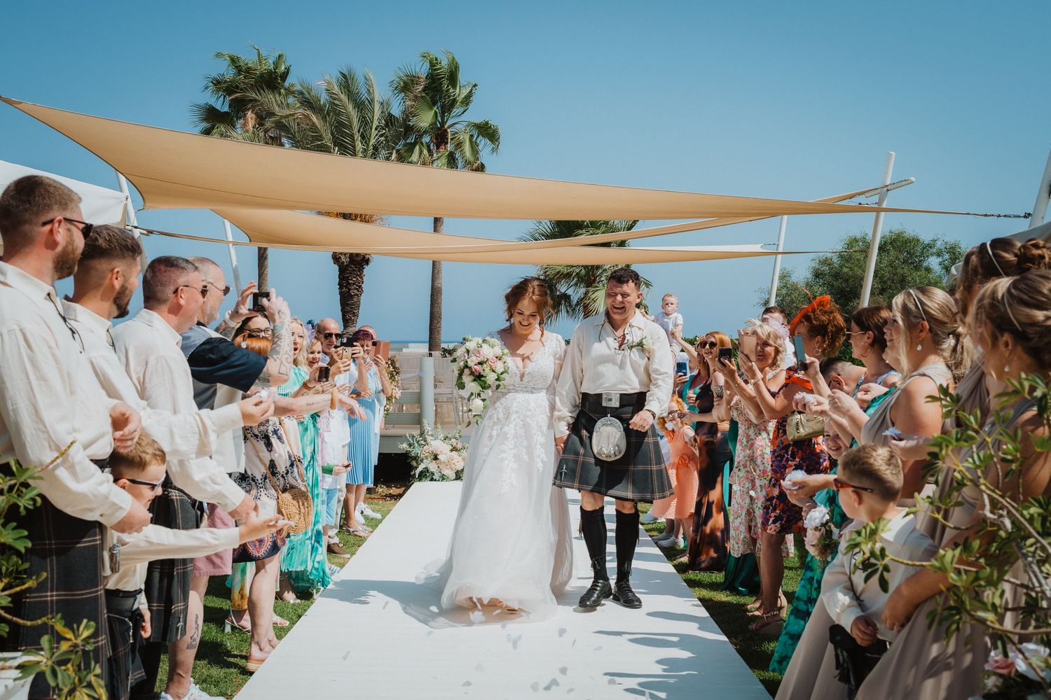 Wedding ceremony taking place outdoors under beige canopy with palm trees and ocean views.