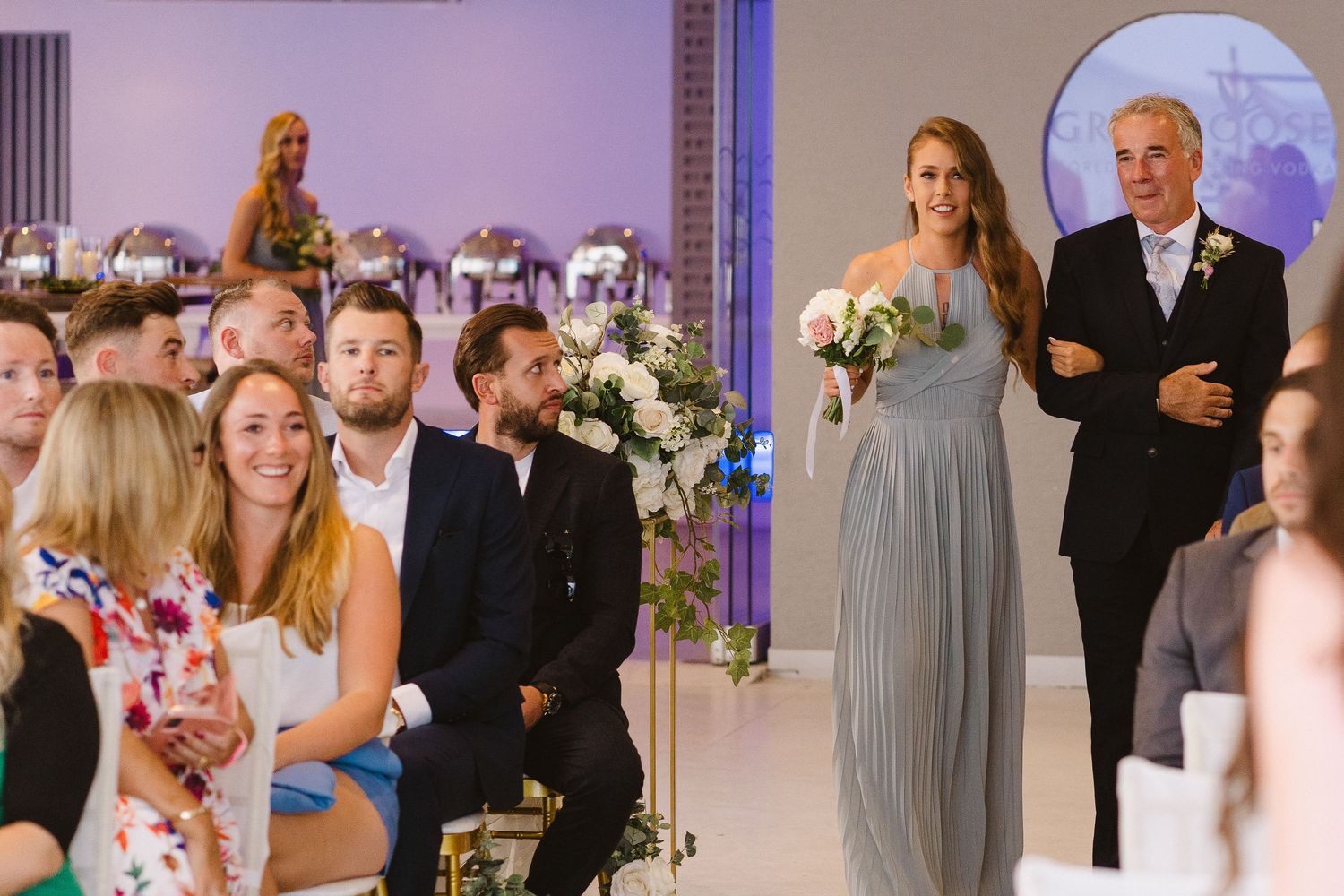 Wedding ceremony procession in an elegant indoor venue with purple accent lighting and floral decorations.