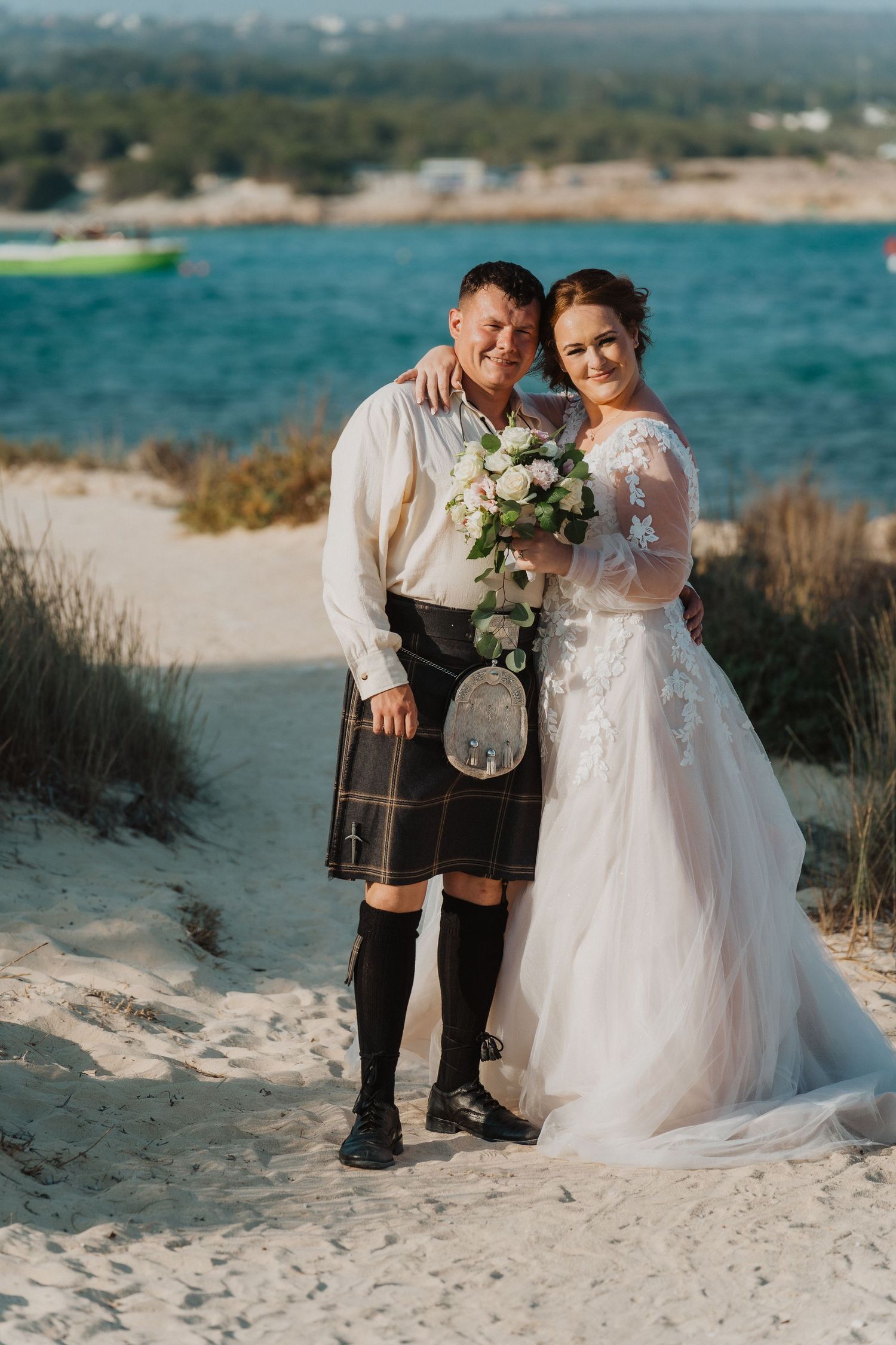 A couple shares a romantic moment on a beach, with the groom in traditional Scottish dress.