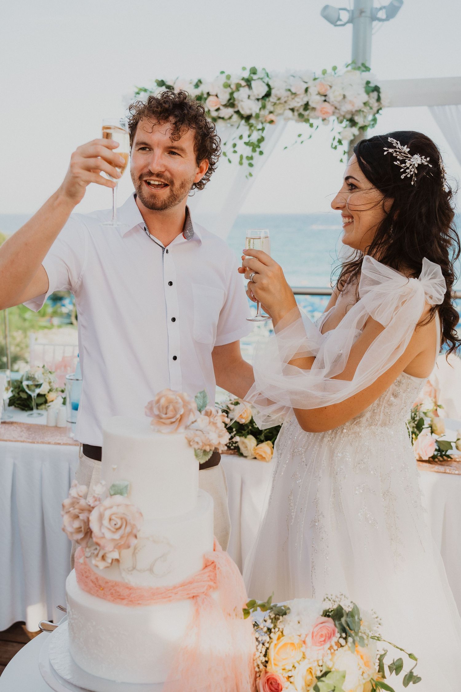 A couple raises their glasses in a toast next to their wedding cake at an oceanfront reception.