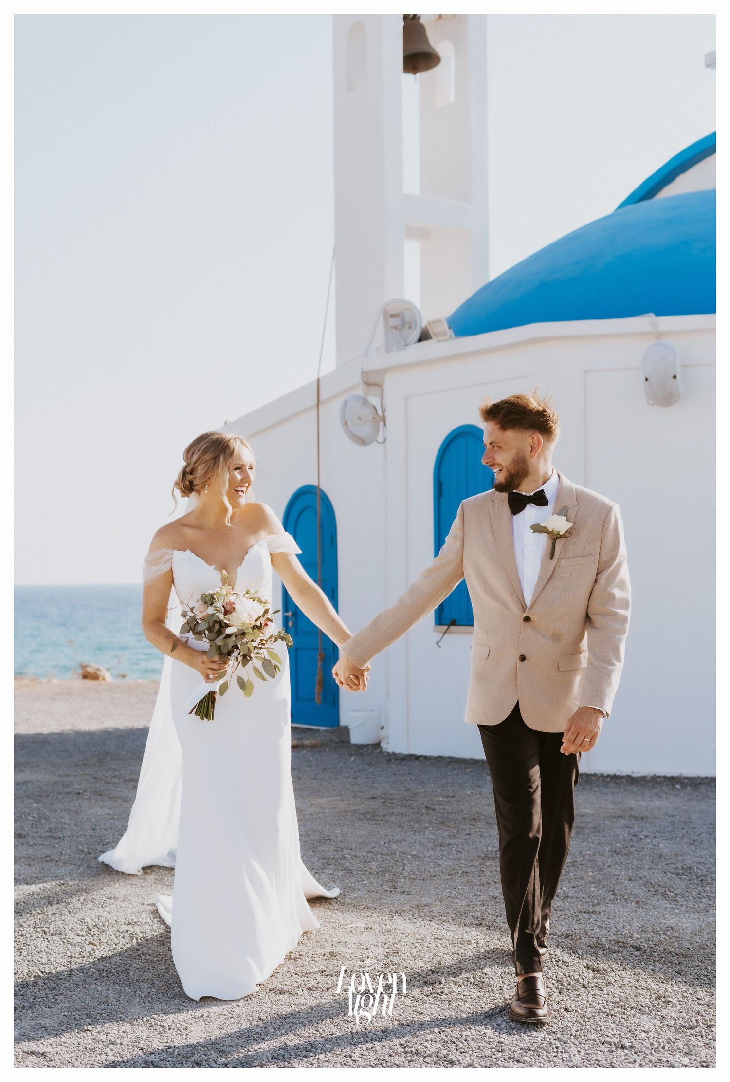 Bride in flowing dress and groom in beige jacket walk hand in hand by seaside Greek church.