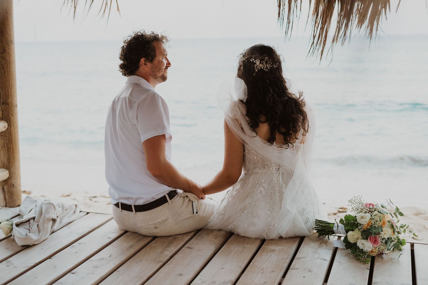 A couple sits together on a wooden deck by the ocean with a wedding bouquet beside them.