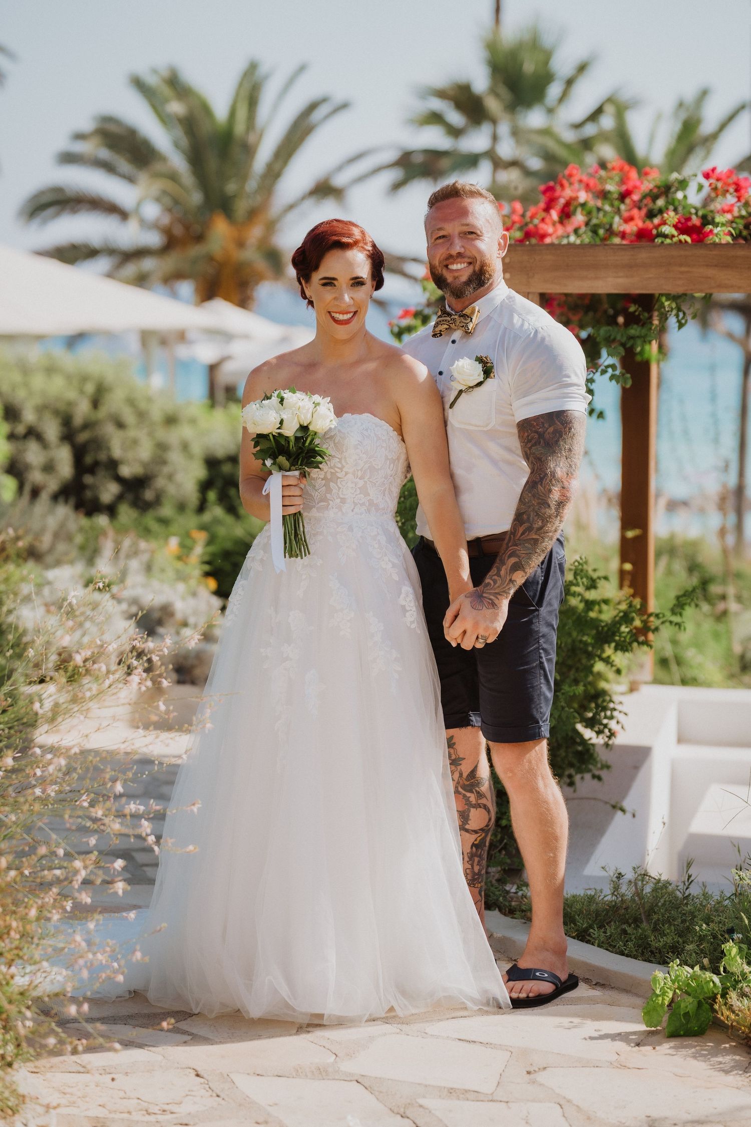 Wedding couple poses in front of tropical garden with palm trees and blooming flowers.