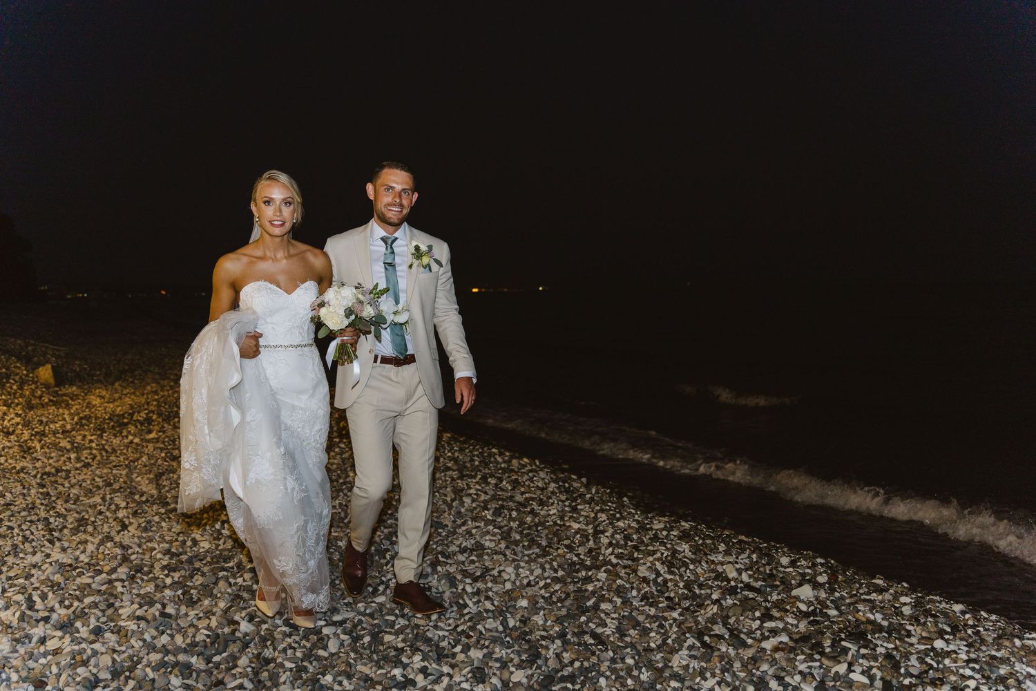Newlyweds share a romantic moment walking together on a pebbled beach in the evening.
