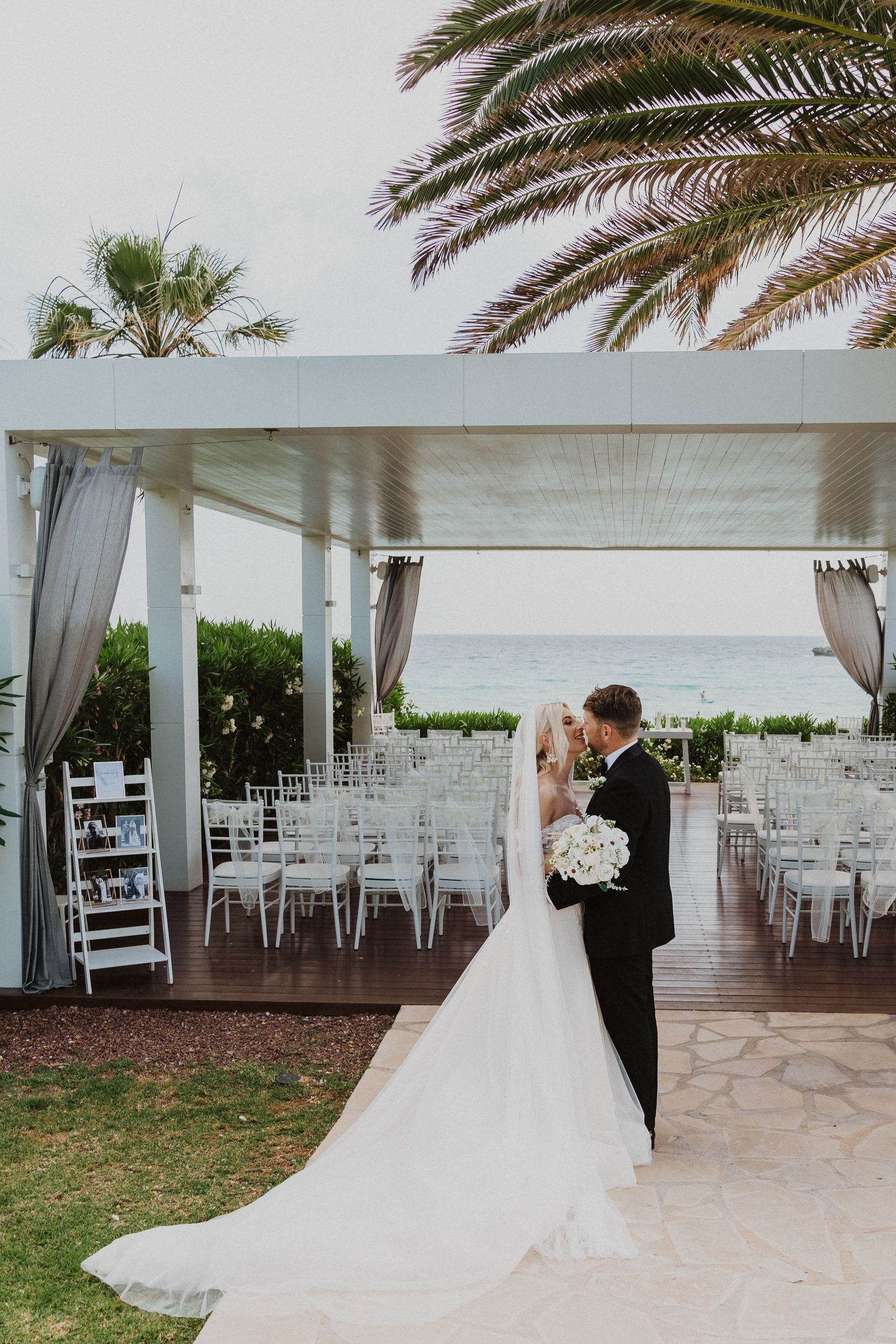 Beachside wedding ceremony setup with palm trees and white chairs under an elegant covered pavilion.