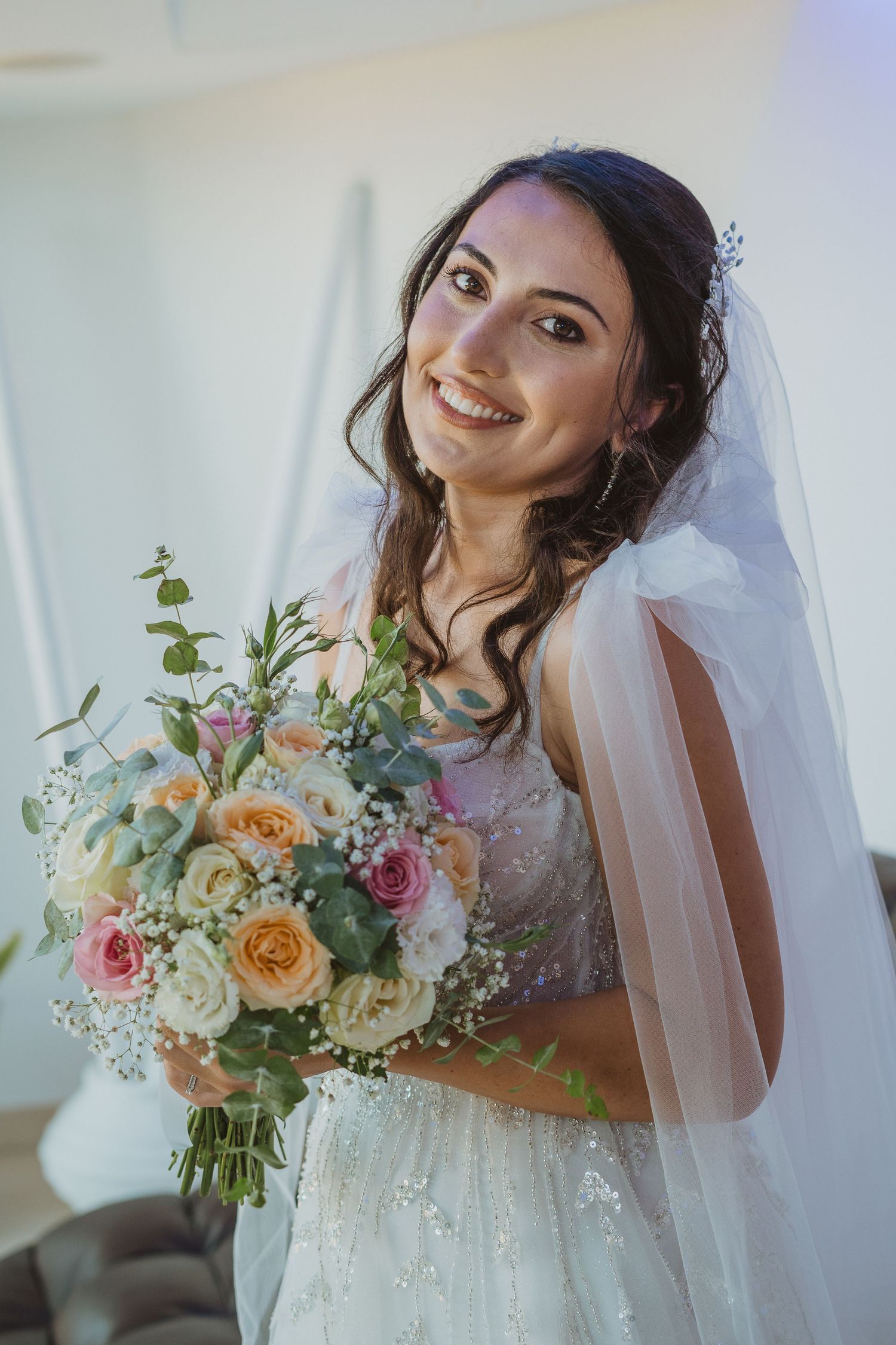Bride wearing lace wedding dress and veil holds beautiful bouquet of pastel roses and mixed flowers on wedding day.