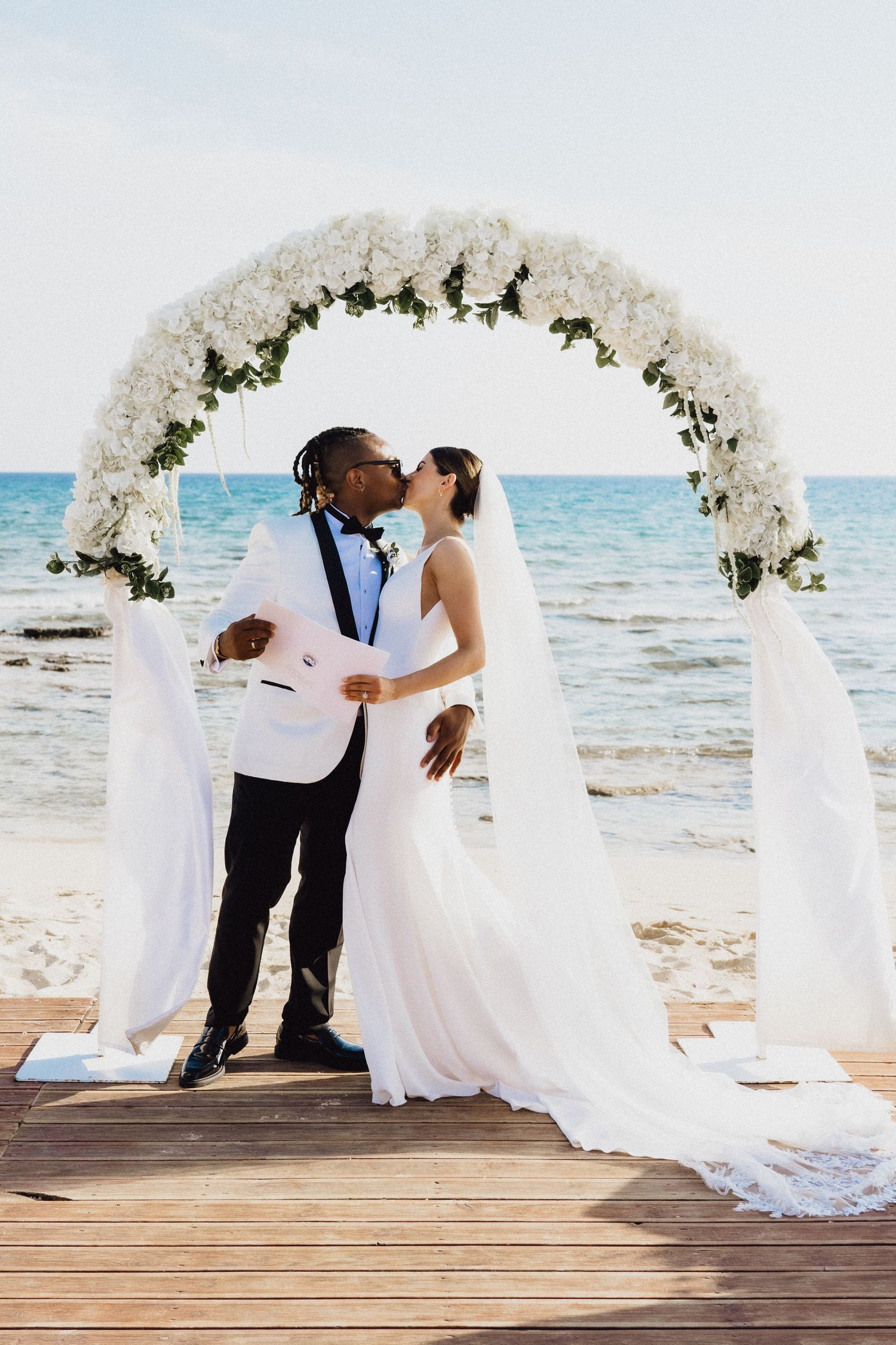 Wedding ceremony under a white floral arch on a seaside boardwalk.