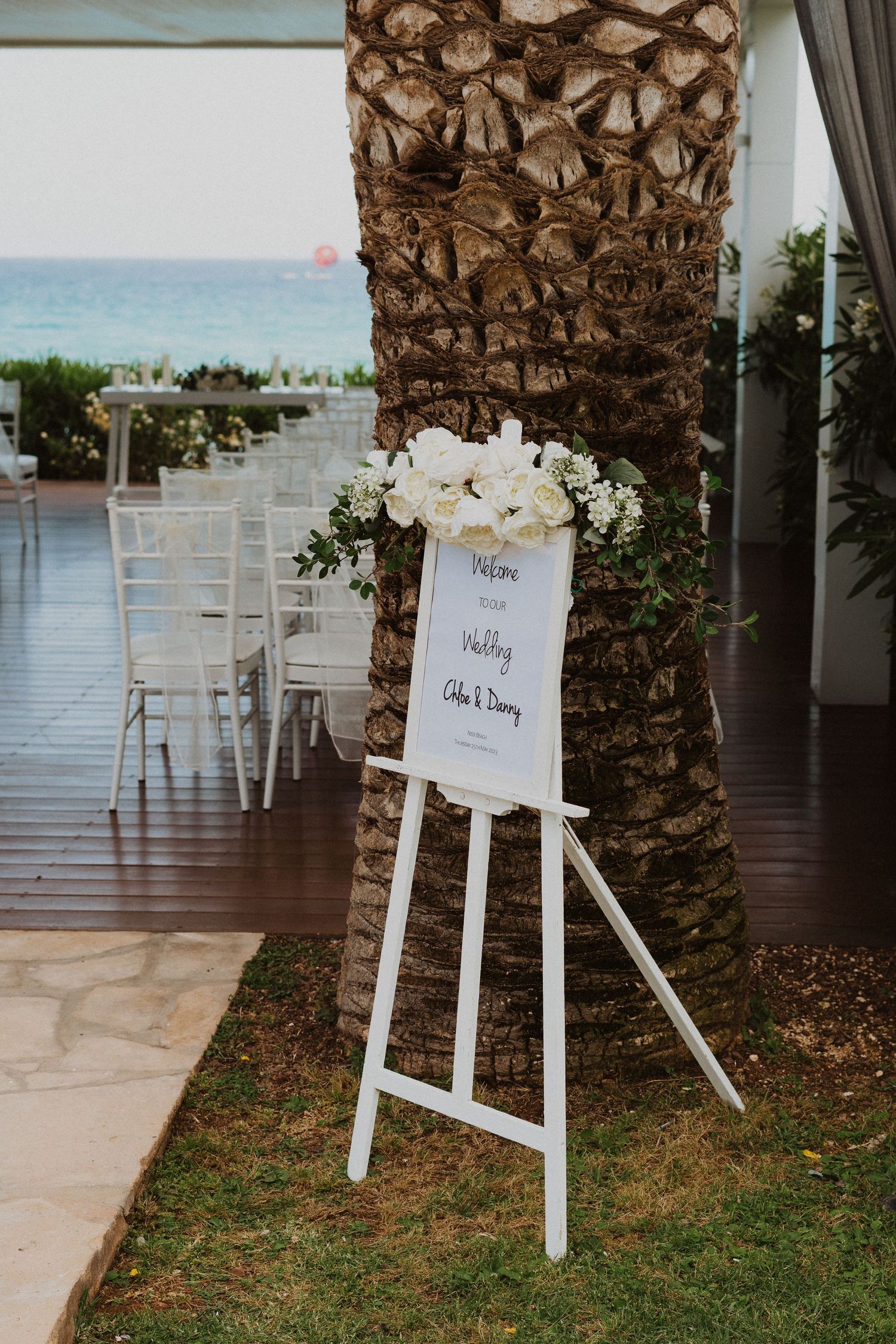 White floral wedding sign on an easel near a palm tree with ocean view in background.