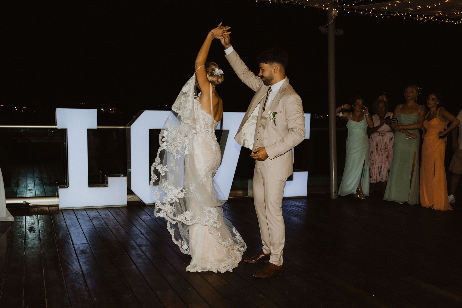 Newlyweds dancing near illuminated LOVE sign at nighttime reception.