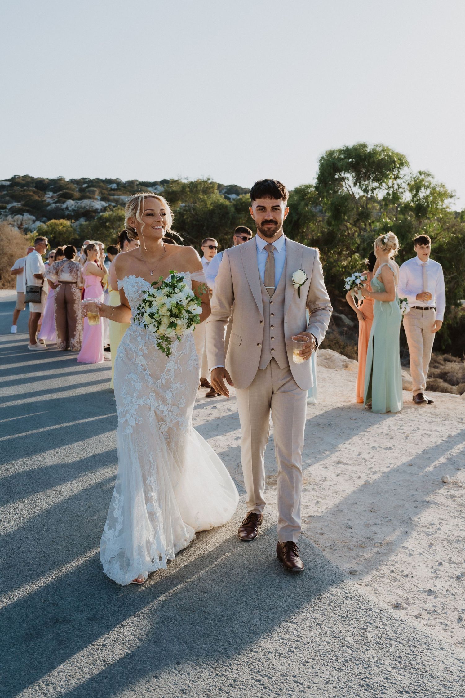 Wedding party members in pastel dresses and light suits walk along a sunlit coastal path.