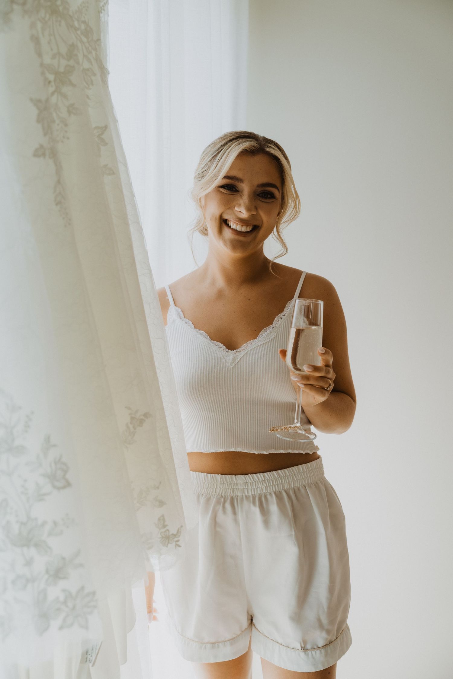 Someone in white loungewear holds champagne glass while standing next to hanging wedding dress.