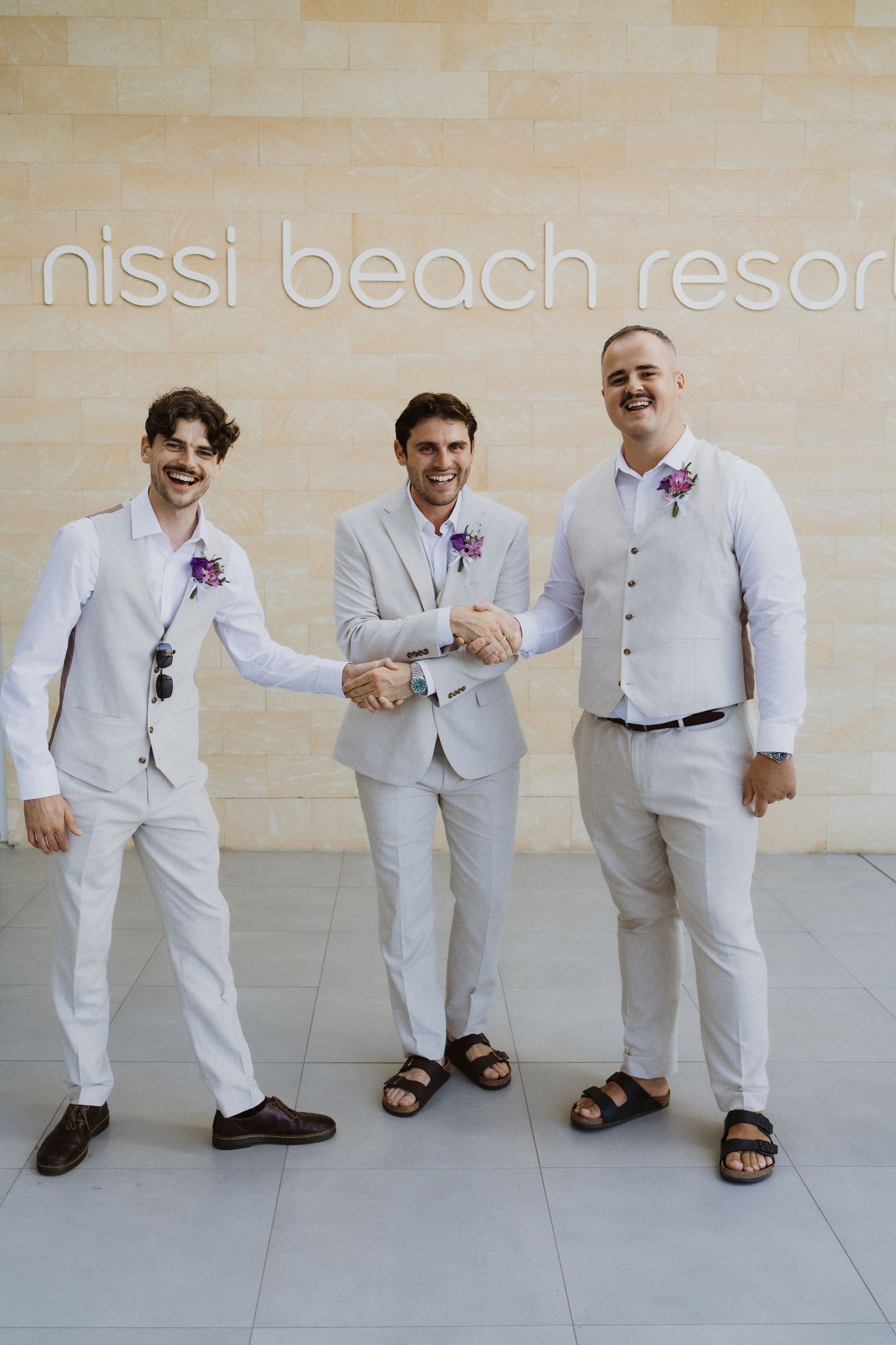 Wedding party in light suits sharing candid moments at Nissi Beach Resort entrance.