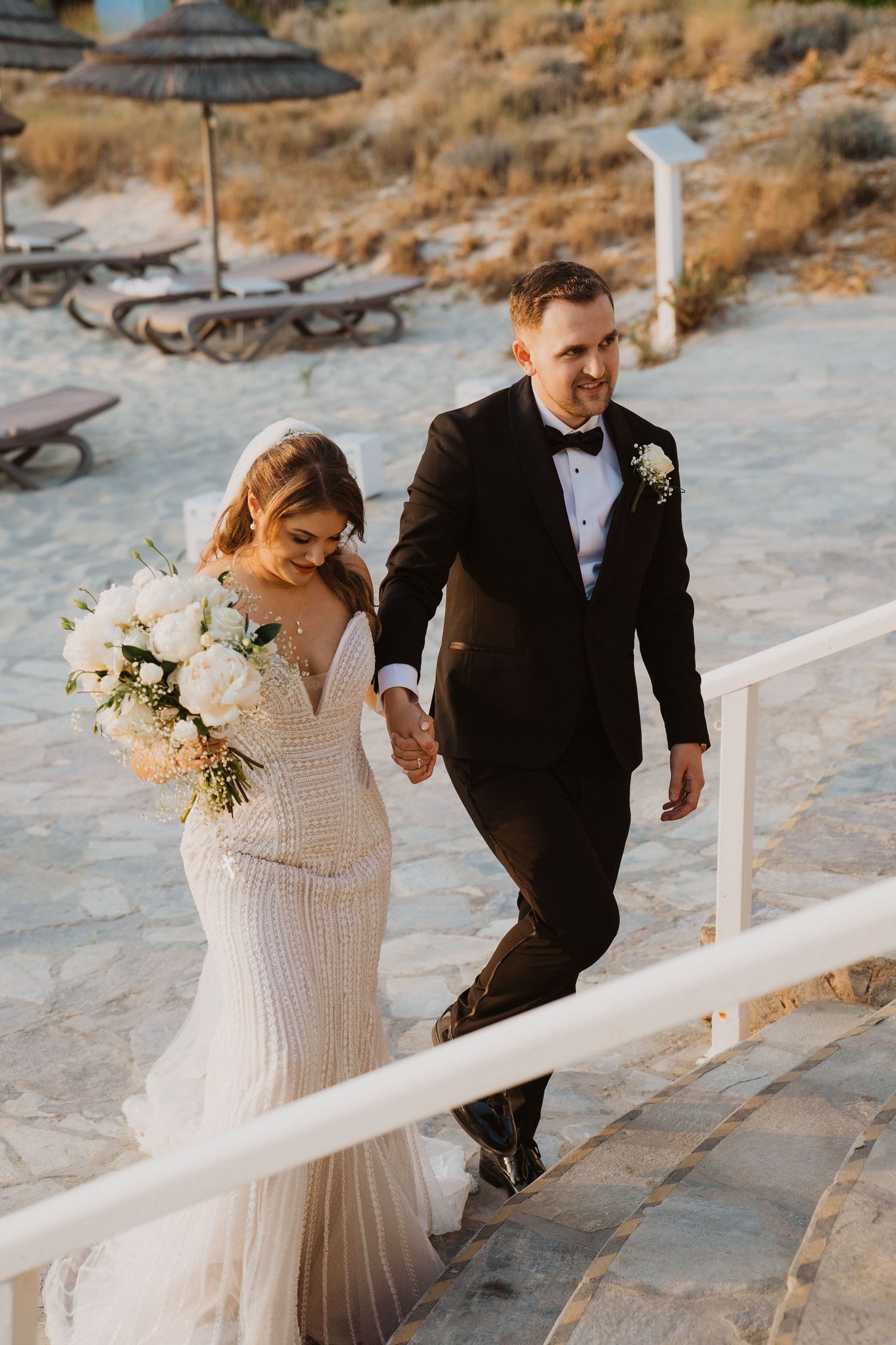 Bride in fitted lace gown and groom in black tuxedo walk hand in hand up steps on a sandy beach.