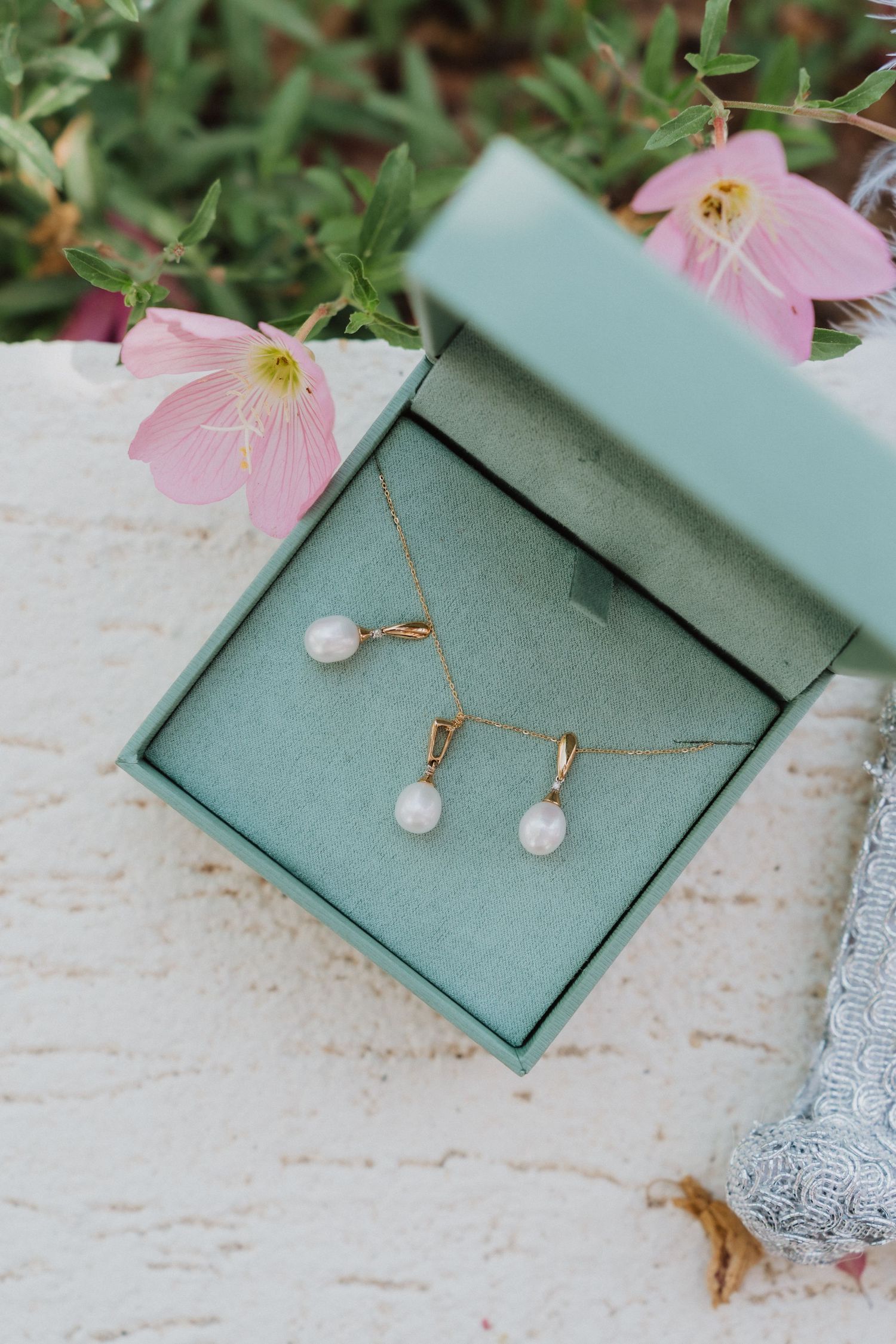 Gold pearl drop necklace and earring set displayed in a teal jewelry box with pink flowers in background.