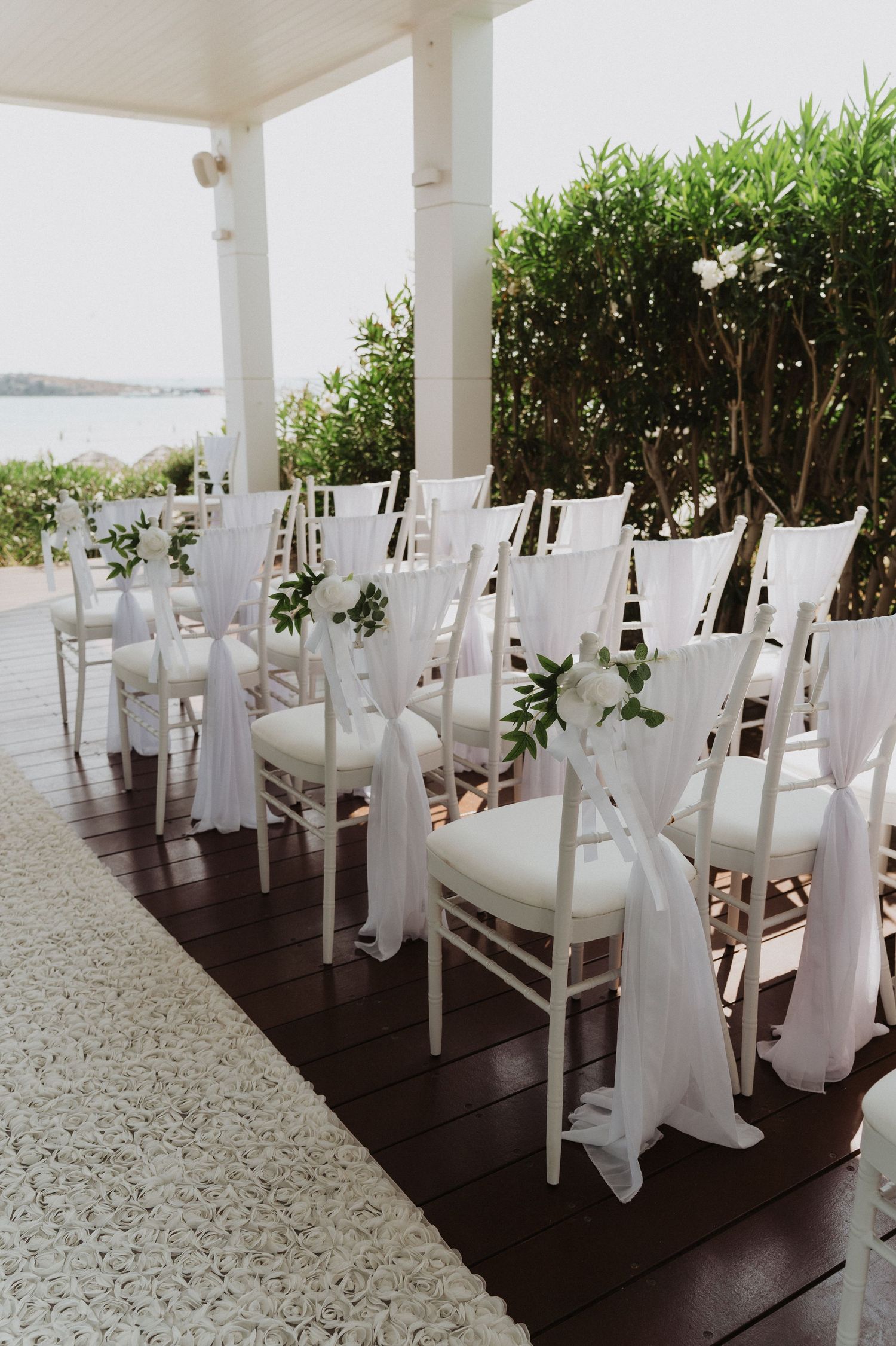 White Chiavari chairs decorated with white fabric sashes and greenery for wedding ceremony.