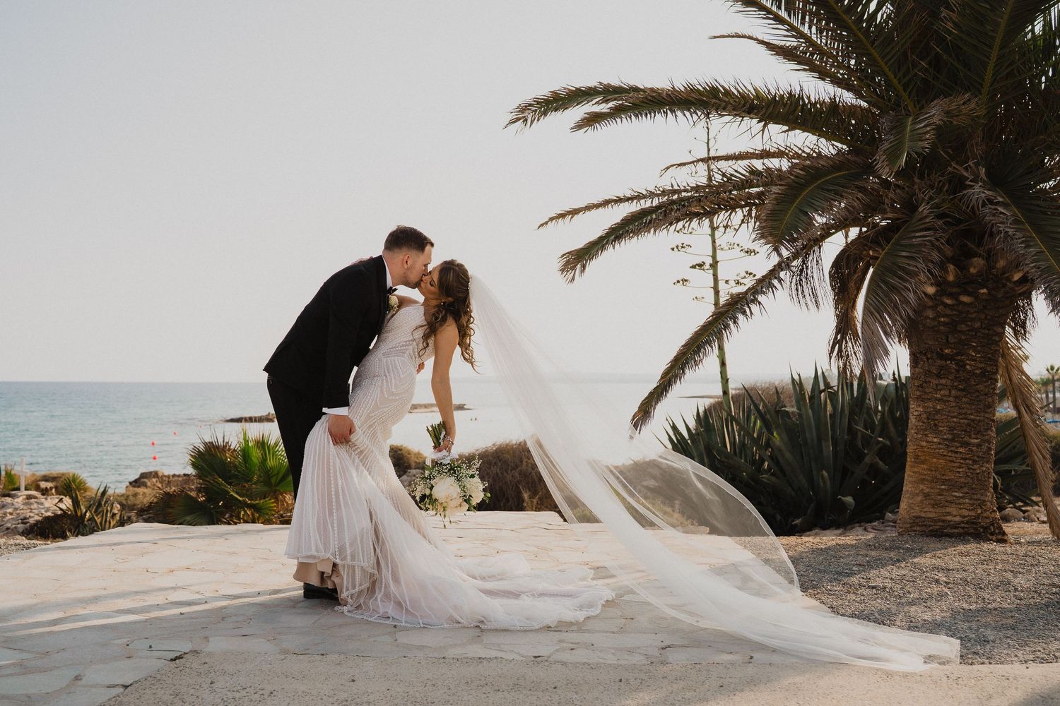 Bride and groom sharing a romantic kiss near a palm tree with the Mediterranean sea in the background.