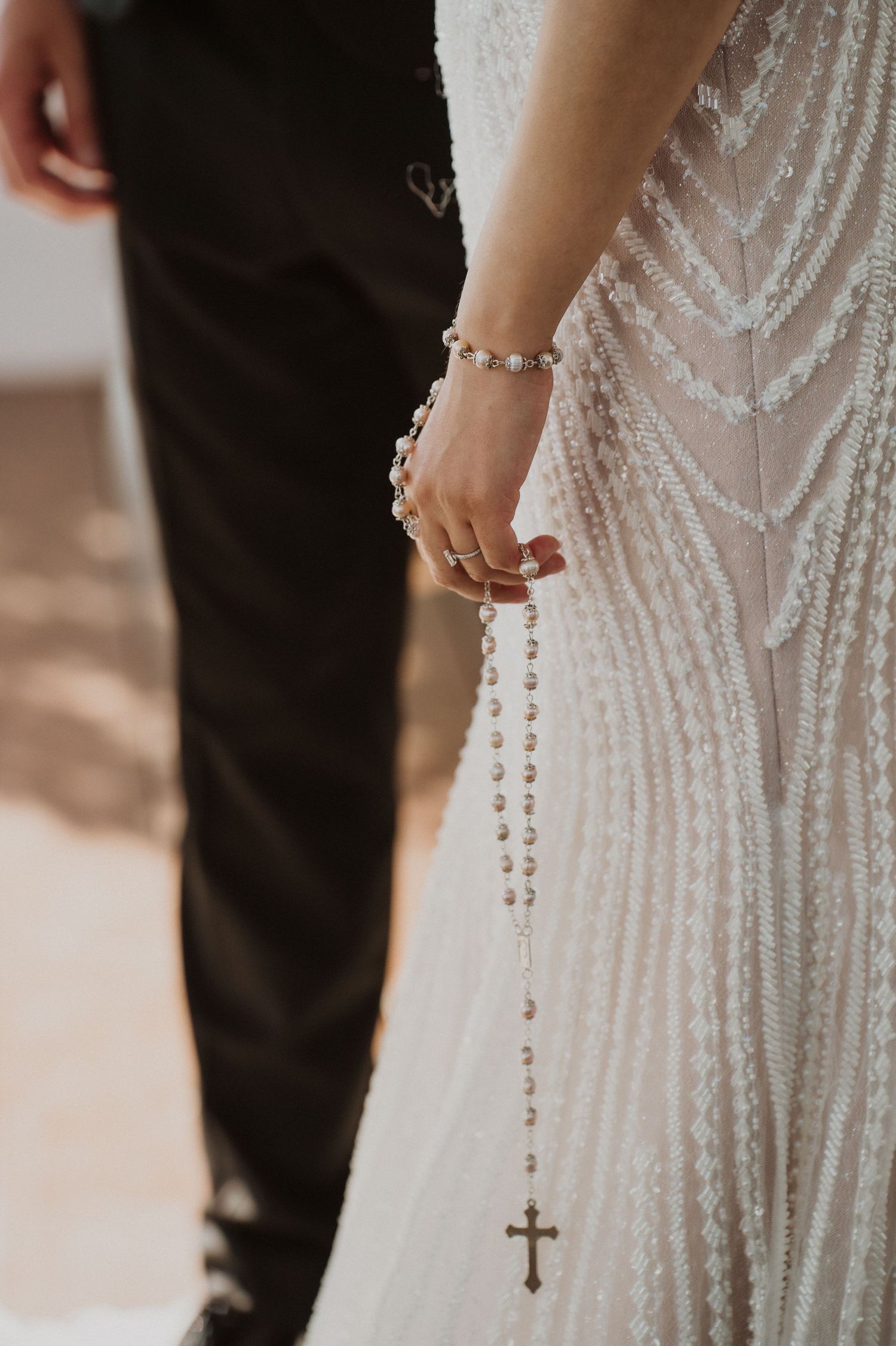 Bride in a beaded white gown holds a pearl rosary with a cross, standing beside a groom in a black suit.