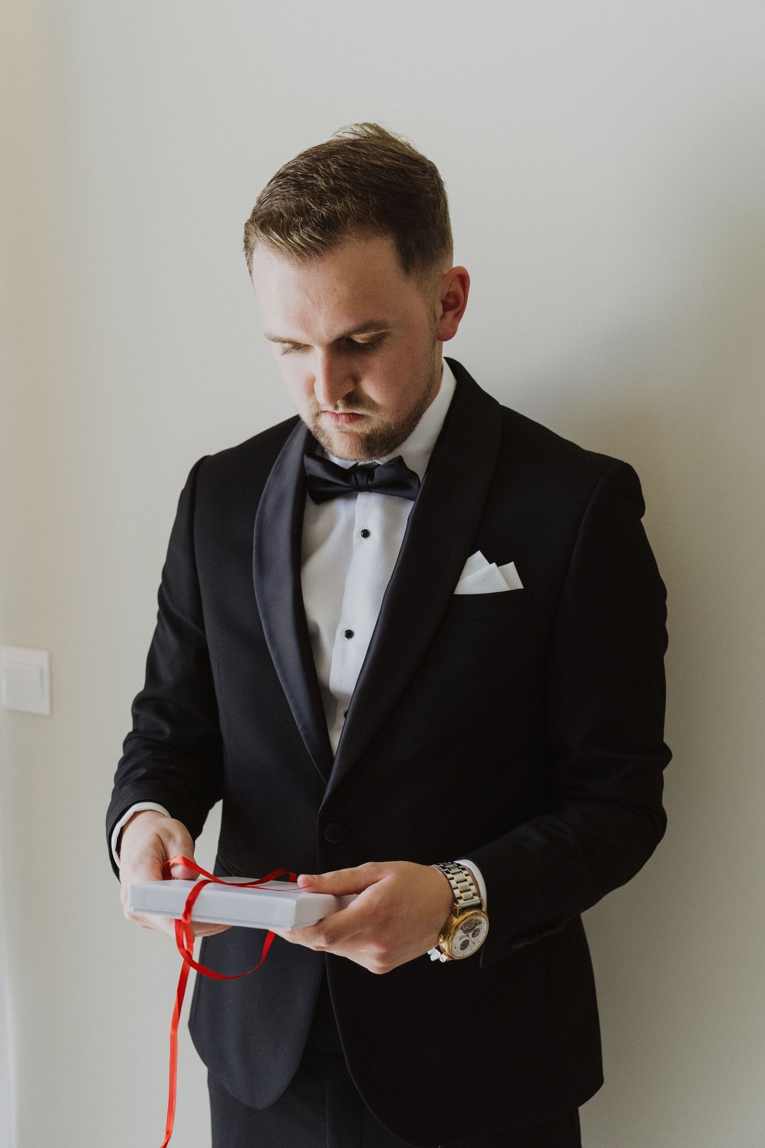 Groom in black tuxedo with bow tie reads a heartfelt letter tied with red ribbon before wedding ceremony.