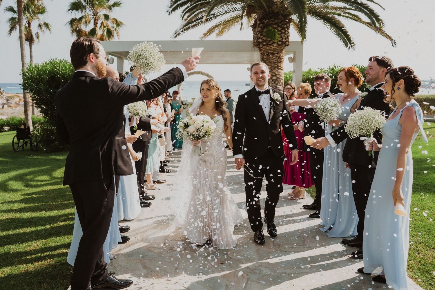 Bride and groom walk down aisle as guests toss confetti at outdoor wedding ceremony near ocean.