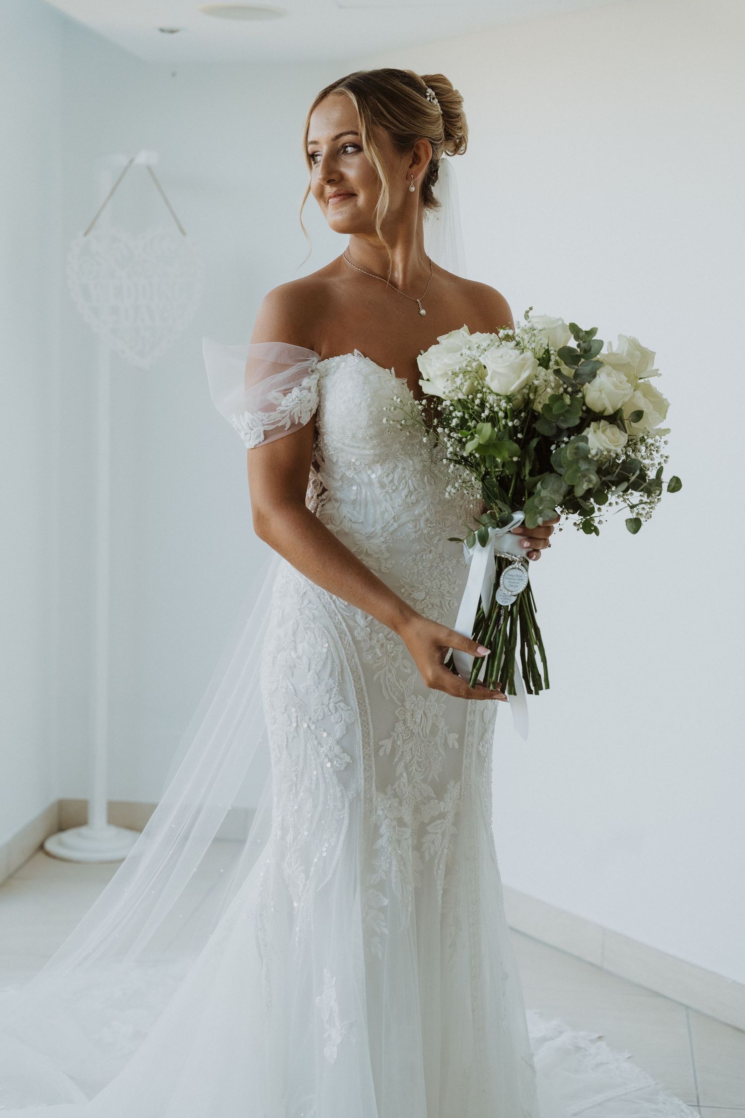 A bride in an off-shoulder lace wedding gown holds a white and green bouquet while standing in a bright room.