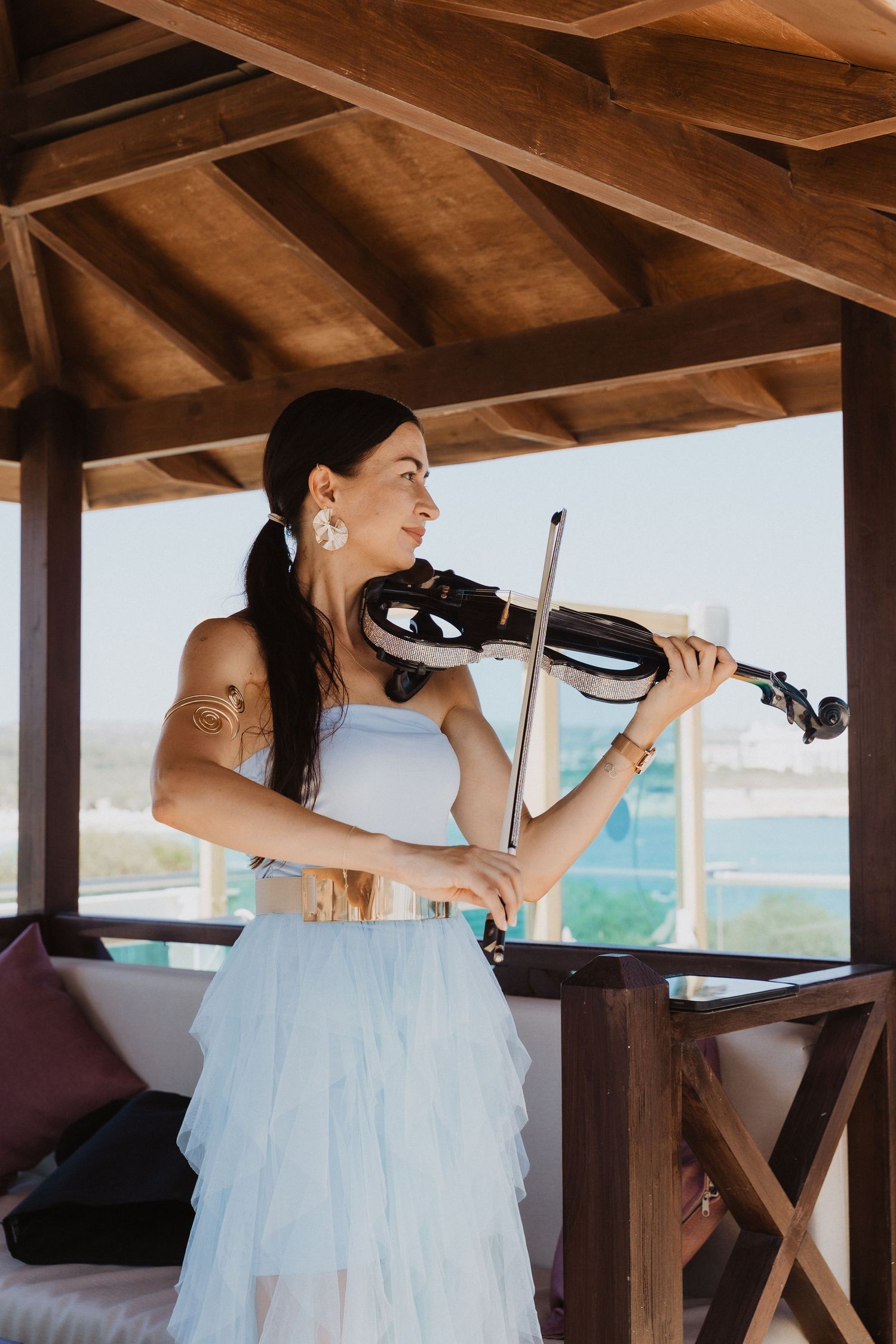 A violinist in a flowing white dress performs under a wooden gazebo with ocean views in the background.