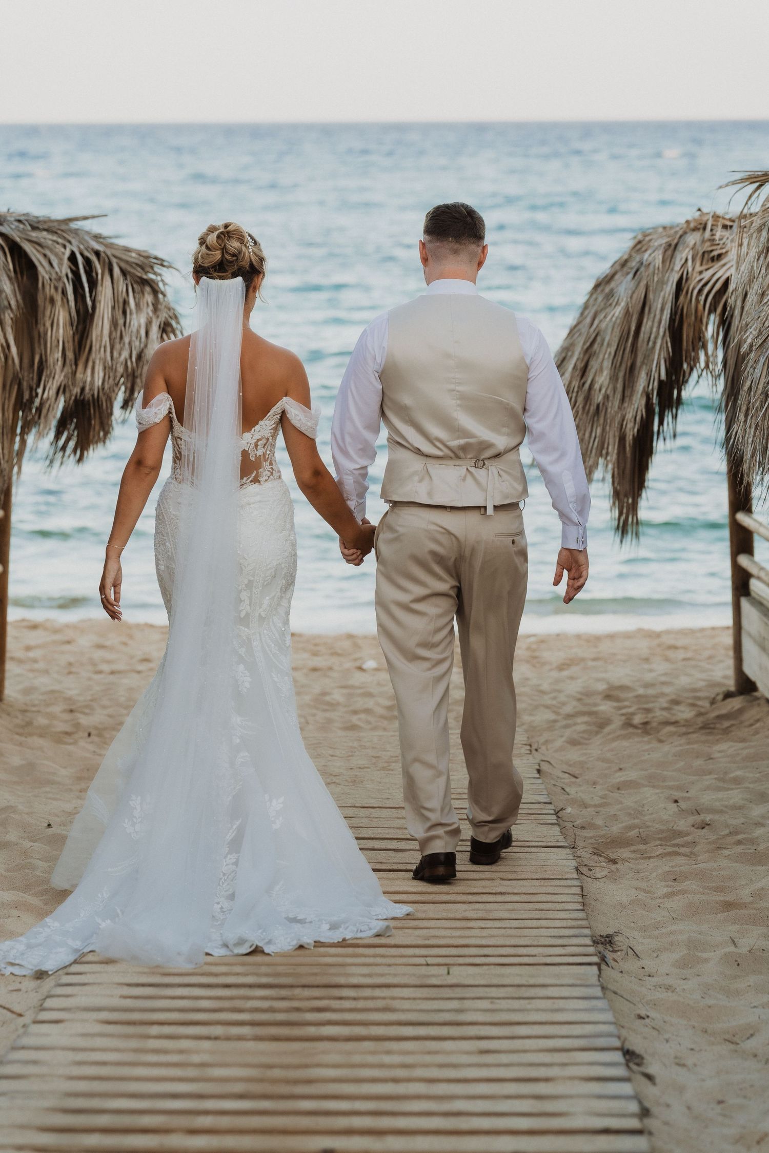 Couple walking down wooden boardwalk on tropical beach with palm frond umbrellas and ocean views.