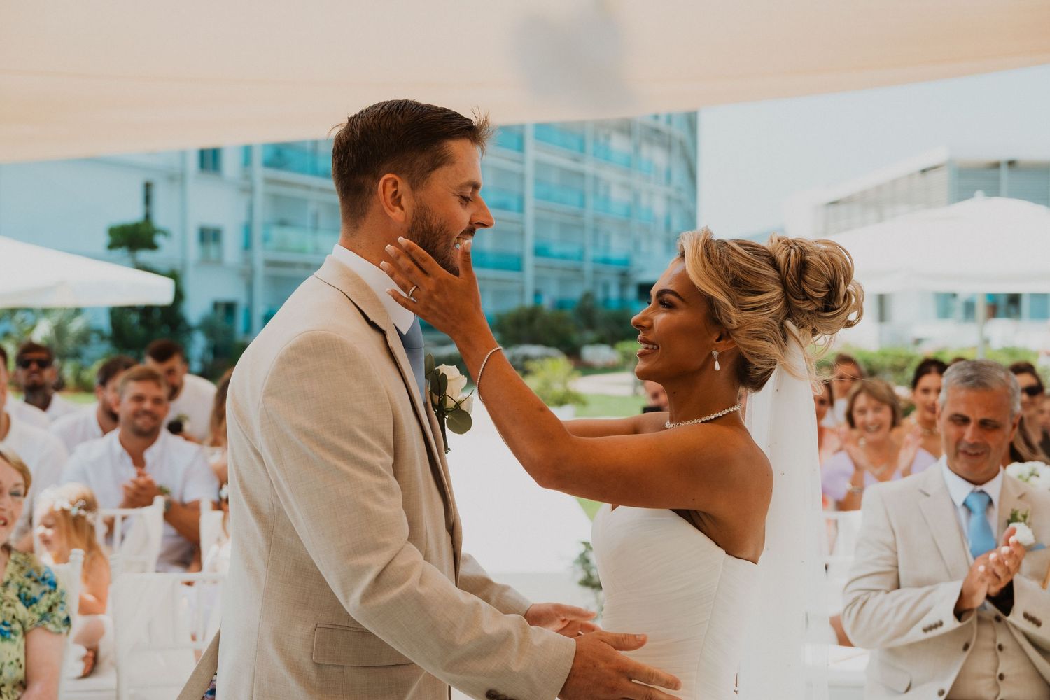 Bride tenderly touches groom's face during romantic first dance at outdoor wedding reception with guests applauding.