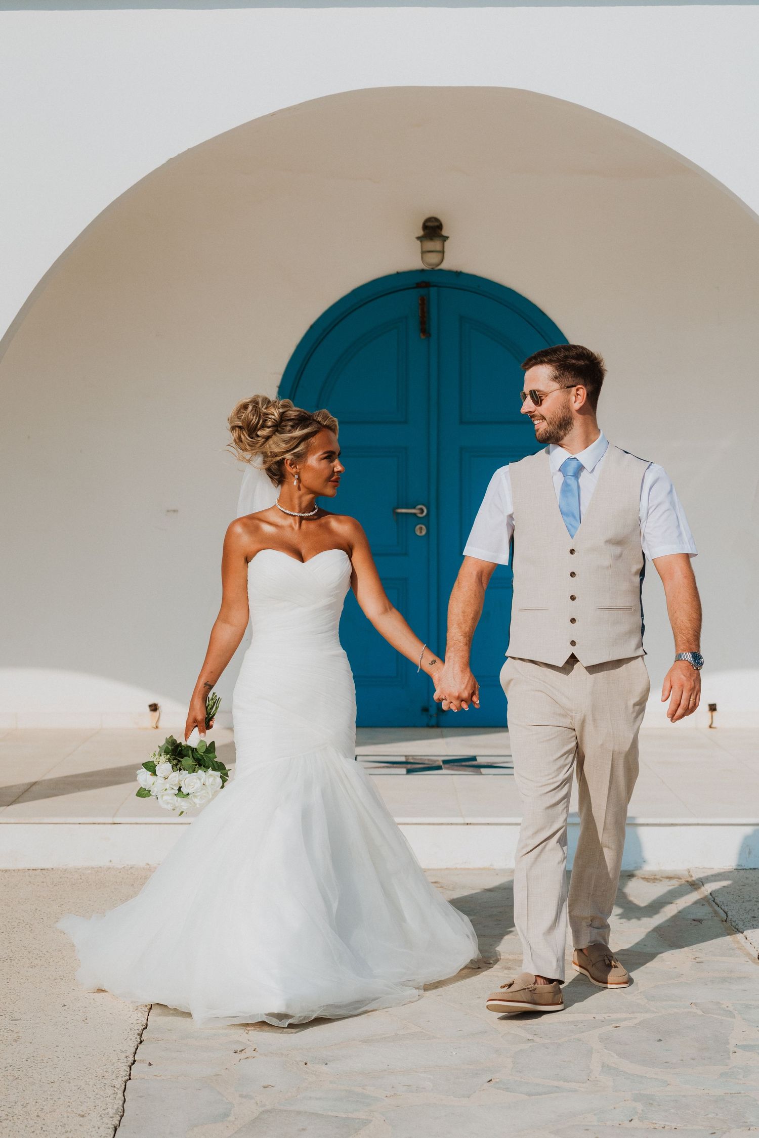 Bride and groom holding hands in front of a white arch with blue door in Santorini, Greece.