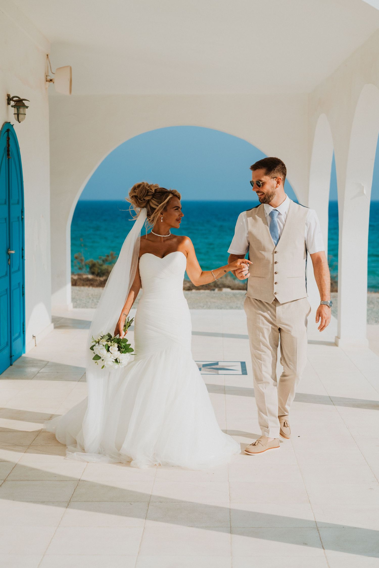 Bride and groom holding hands at a Mediterranean wedding venue with white arches and turquoise sea backdrop.