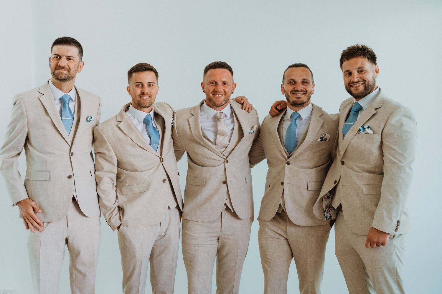 Groomsmen wearing matching beige suits with light blue ties stand together smiling in a formal wedding party photo.