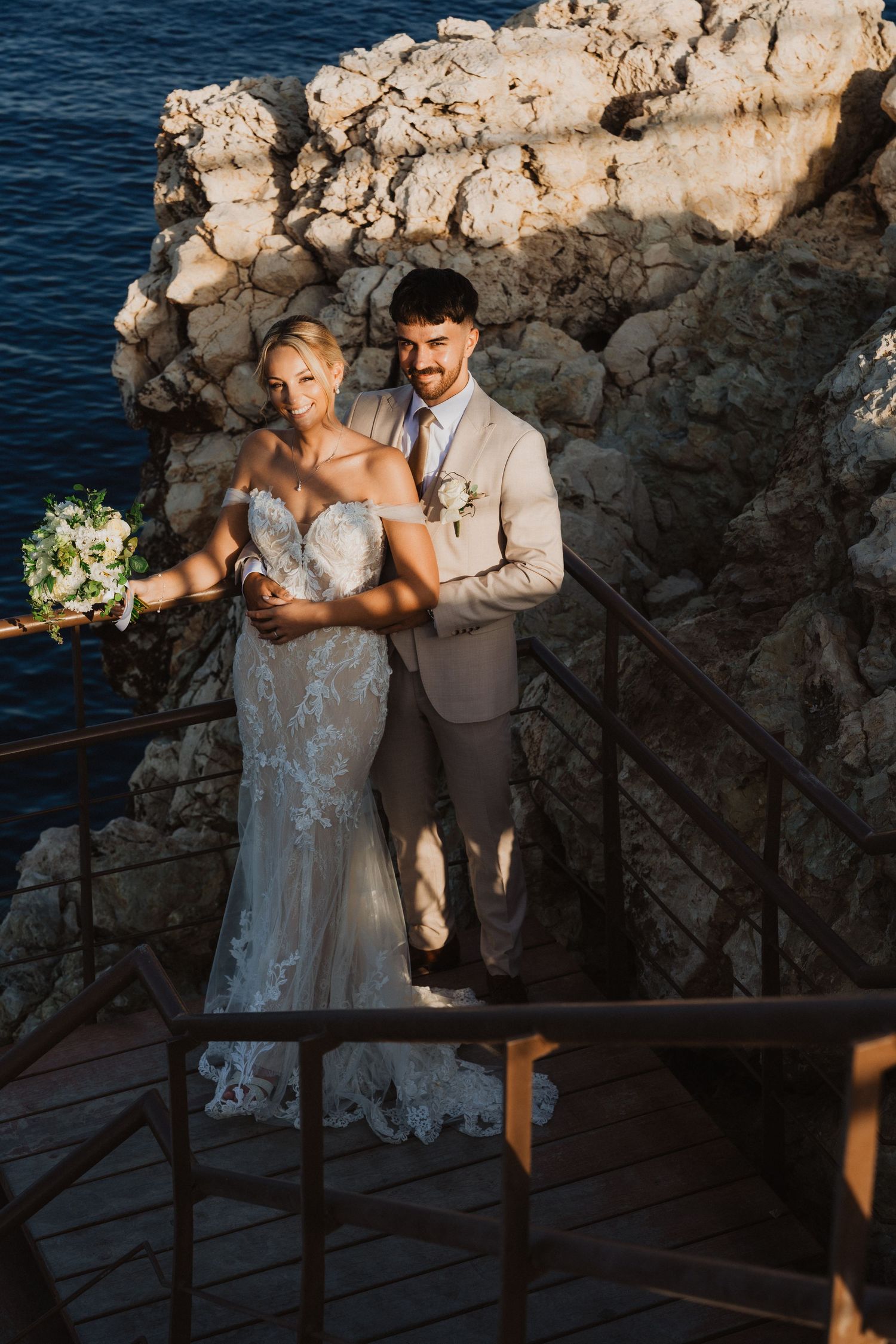 Wedding couple posing on rocky coastal overlook with white bouquet and beige attire at sunset.