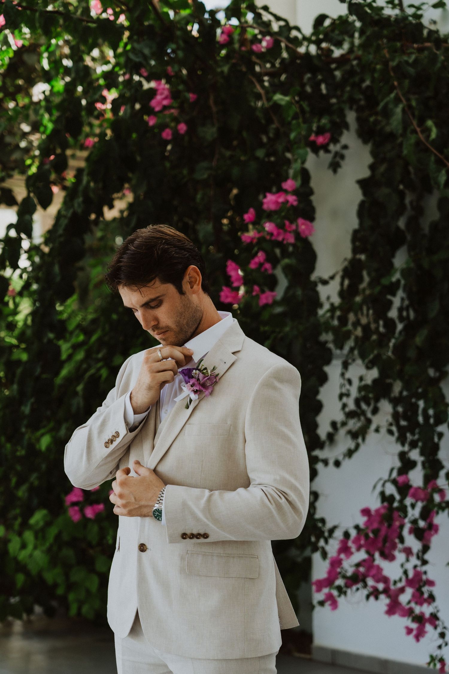 Groom in cream suit shows emotion during wedding moment against floral backdrop.