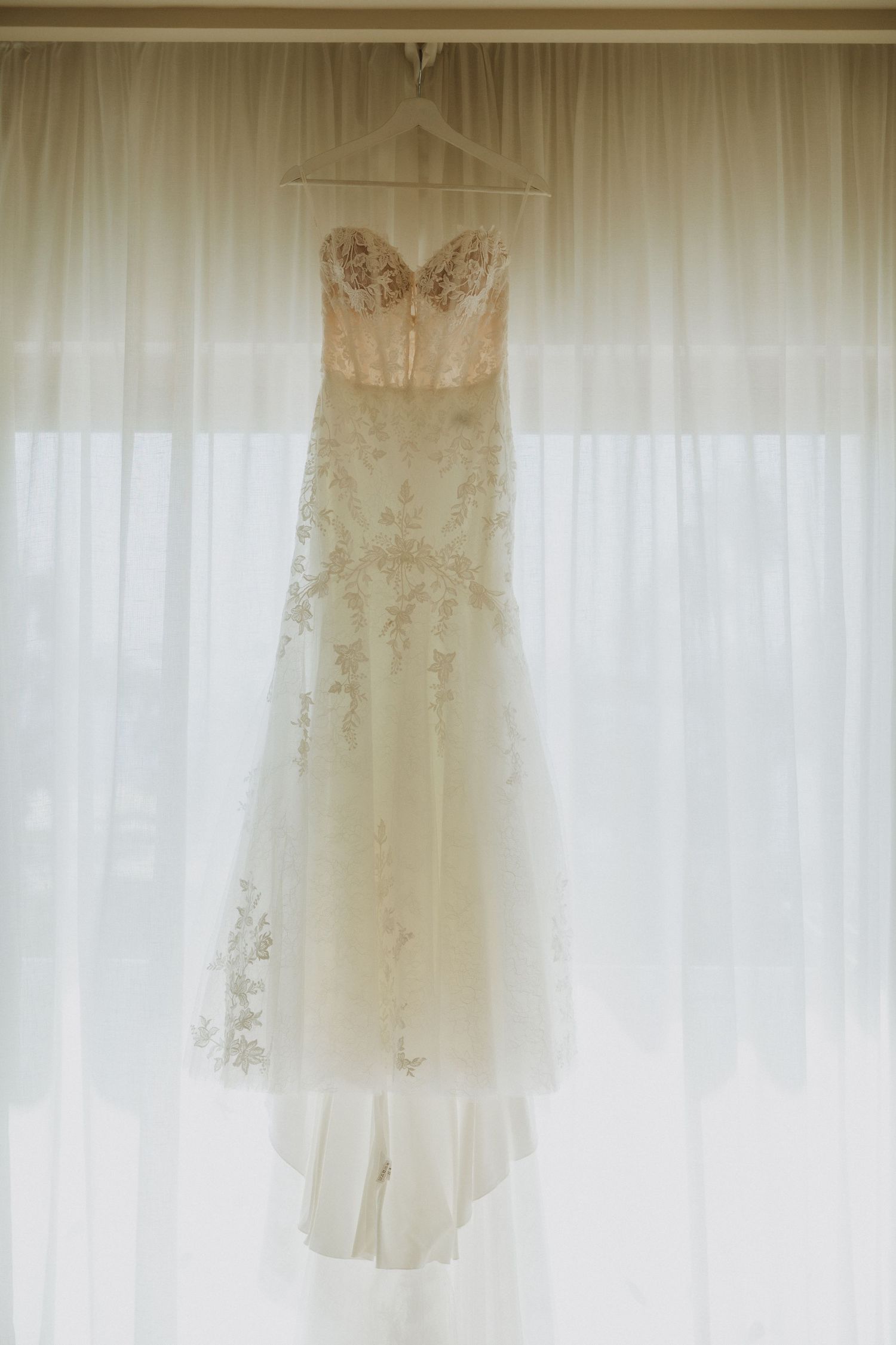 An elegant white wedding dress with lace details hangs against sheer curtains in natural light.