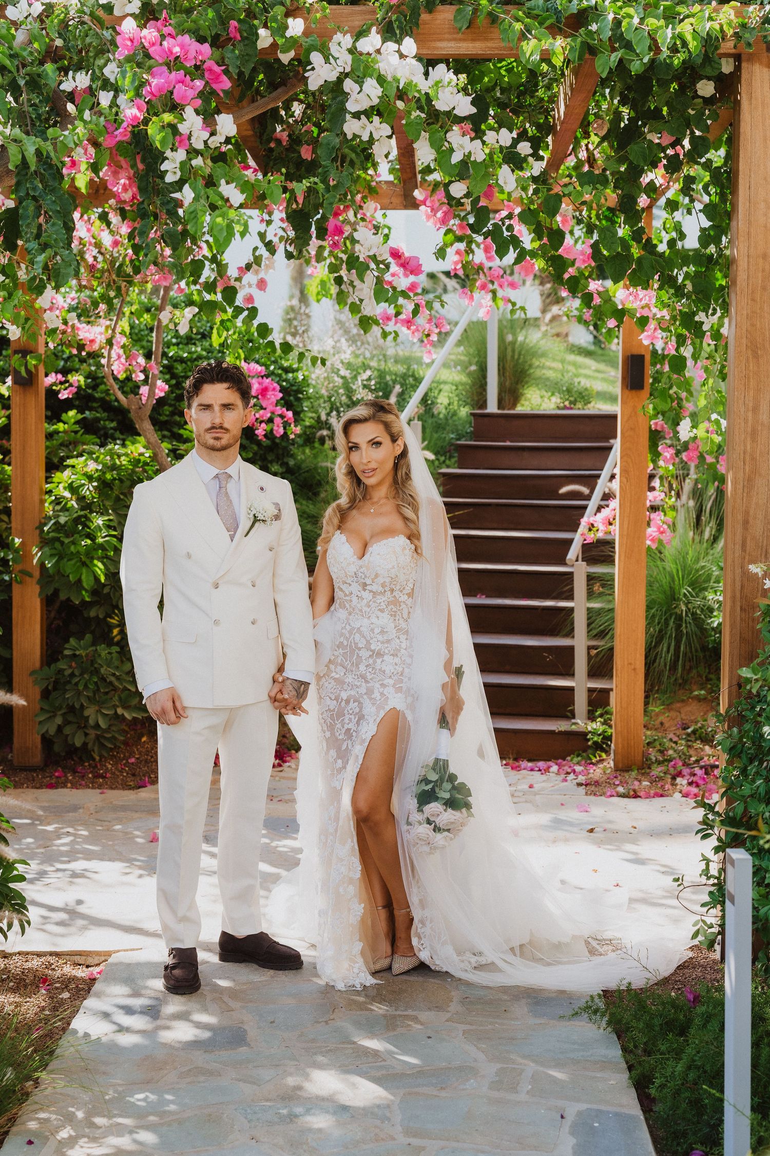 A couple poses under a romantic garden pergola decorated with pink and white flowers on their wedding day.