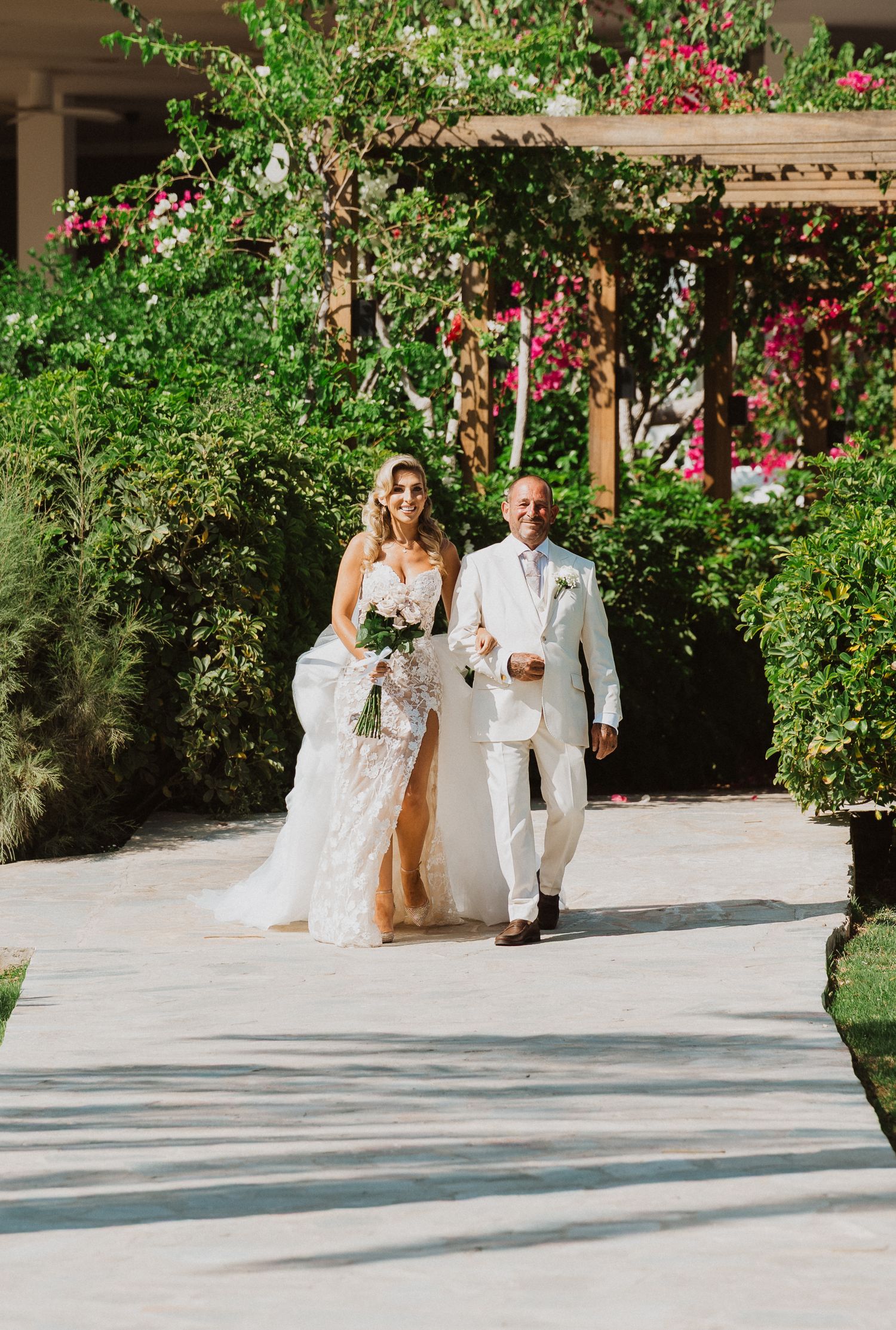 A couple walks down a garden path during a wedding ceremony with flowering bushes and a pergola in the background.