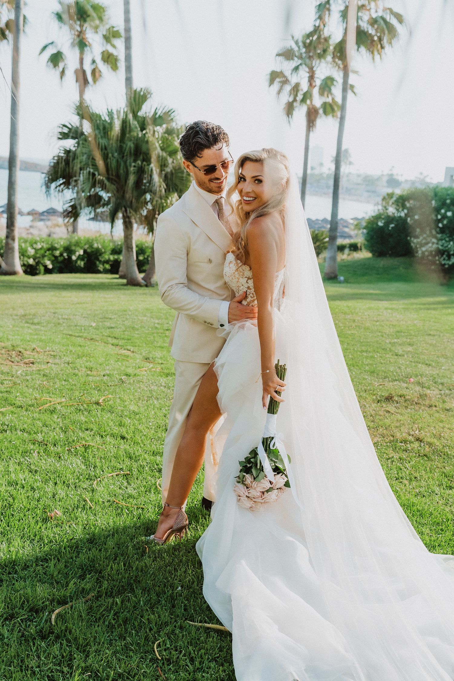 A romantic wedding couple embraces on a sunny lawn with palm trees and ocean views in the background.