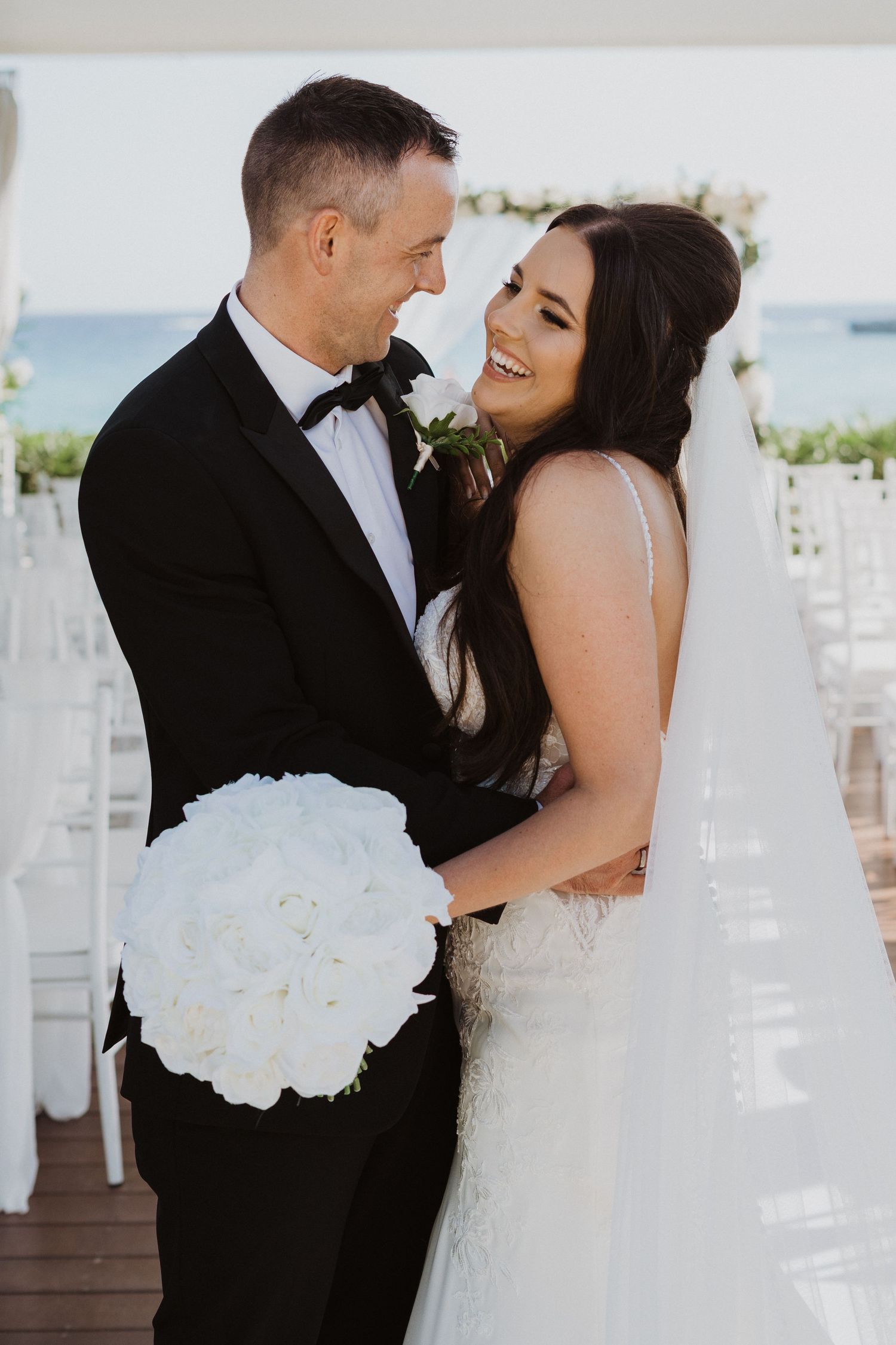 Romantic wedding couple embracing at seaside venue with white floral bouquet.
