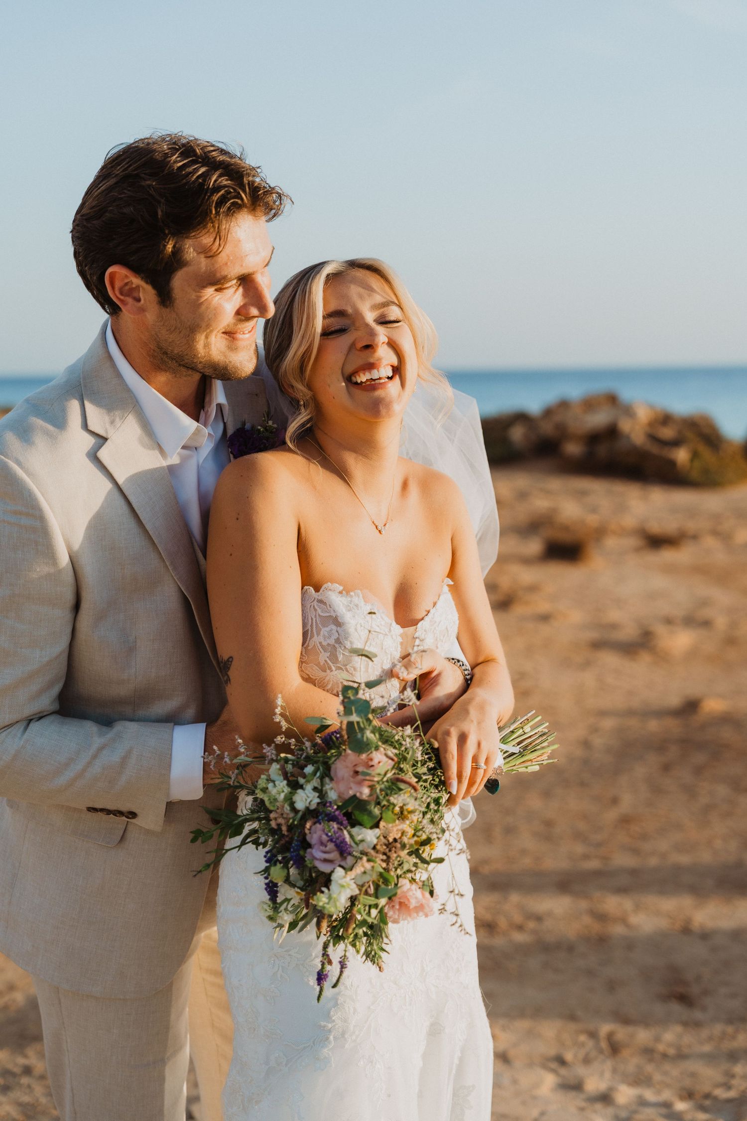 Newlyweds share intimate moments on a rocky beach at sunset while holding a beautiful floral bouquet.
