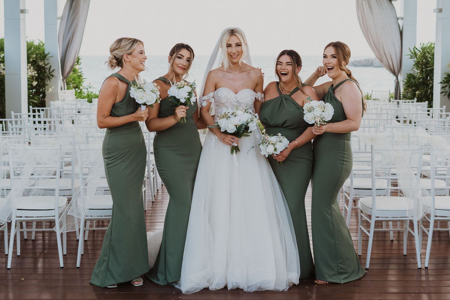 Bridal party in sage green dresses poses with bouquets in front of white ceremony chairs.