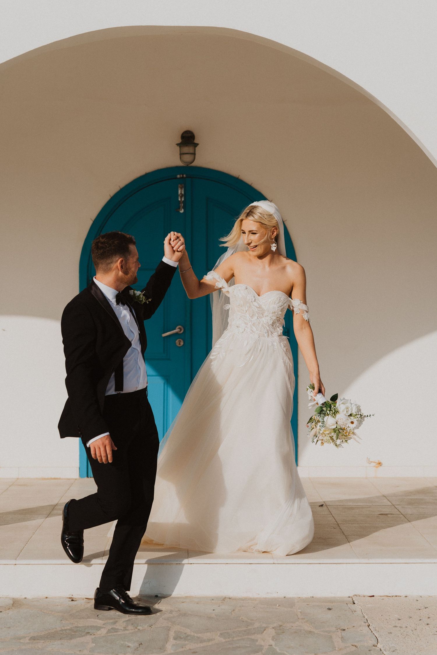 Joyful wedding couple dances in front of turquoise door under white Mediterranean archway.