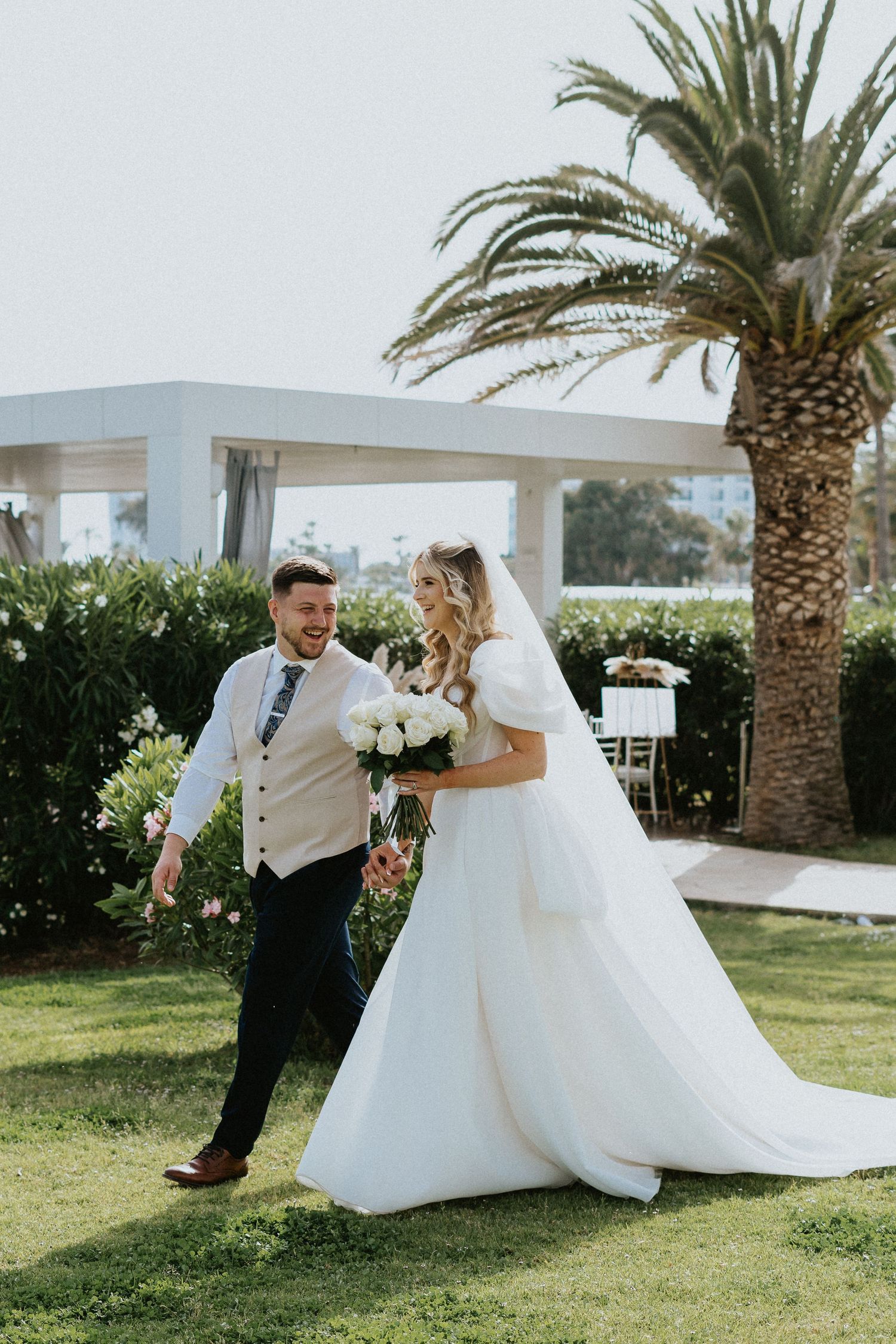 Wedding couple walking on grass lawn with palm tree and white building in background at outdoor venue.