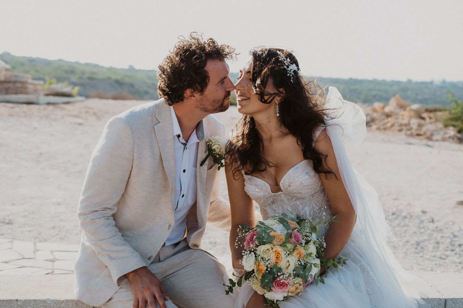 A tender moment between newlyweds against a coastal backdrop with a beautiful bouquet.