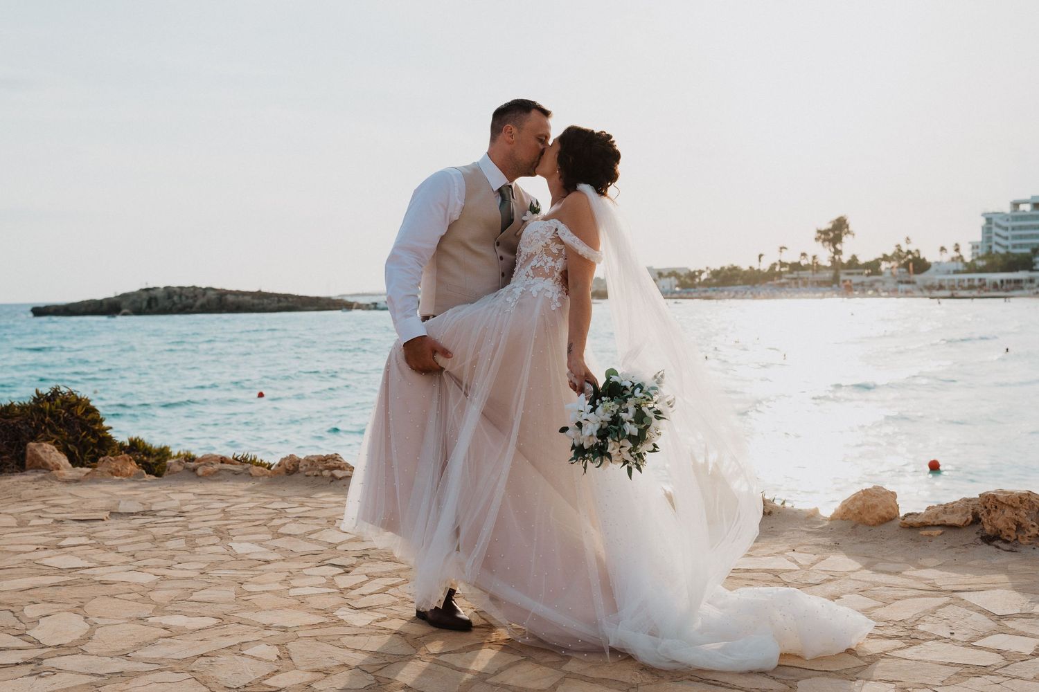 A romantic couple sharing a tender moment on a coastal stone path during their sunset beach wedding ceremony.